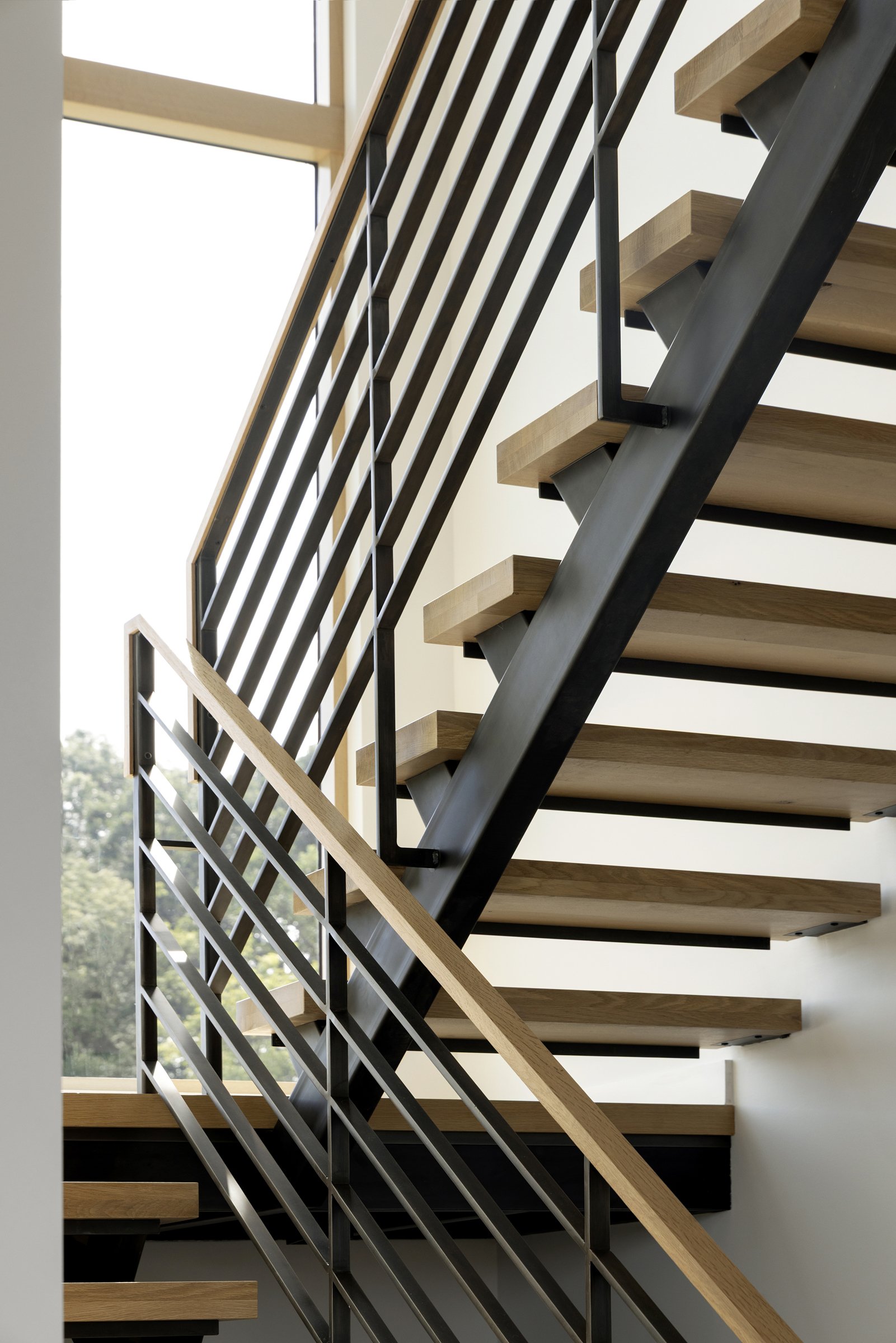 Modern black steel horizontal bar railing with a warm wood handrail by Red Iron Studio, paired with a floating wood tread staircase and central steel stringer in a bright, minimalist interior.