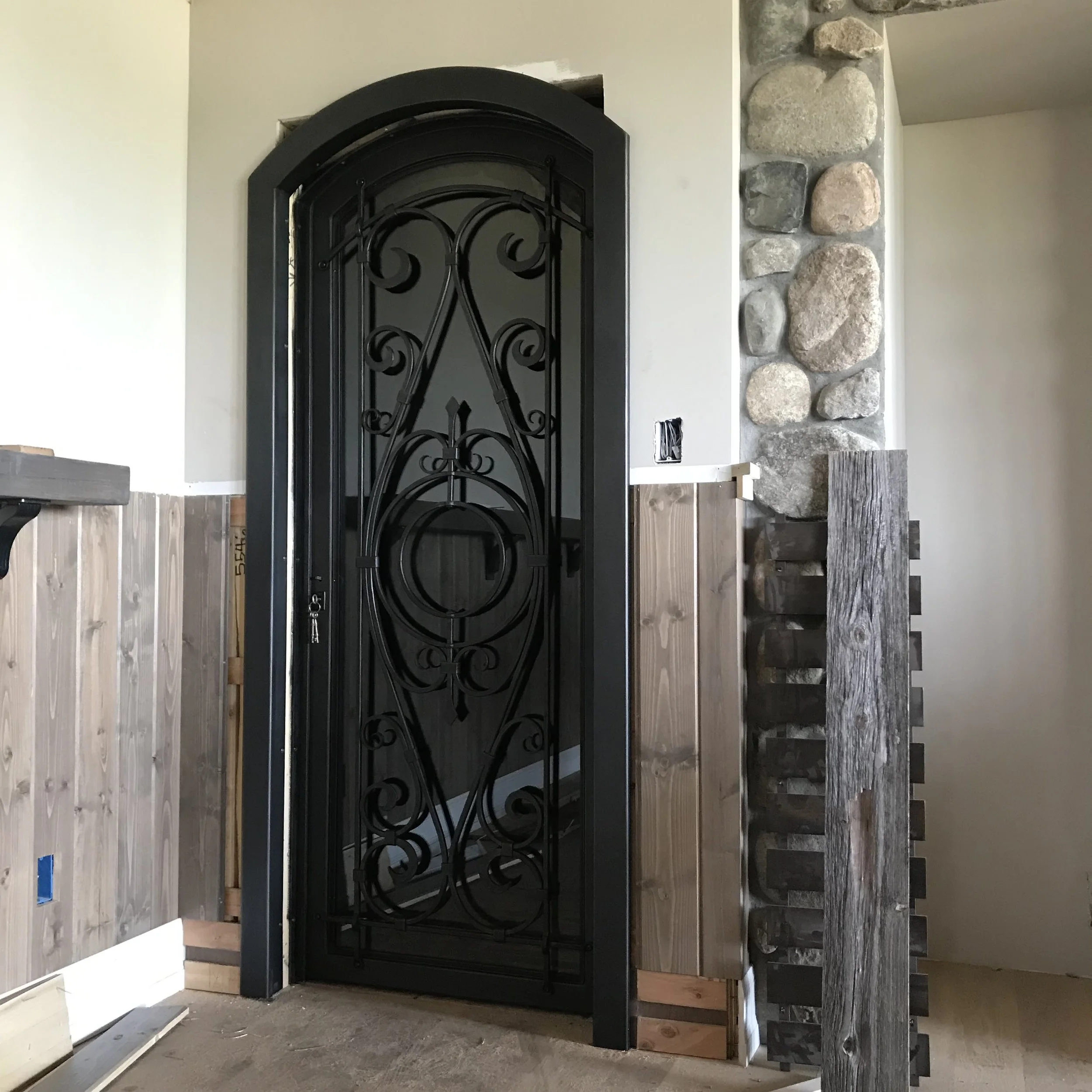 Custom Wrought Iron Wine Cellar Door with Arched Frame - This custom wrought iron door features elegant scrollwork and a decorative circular medallion set within a black arched frame, glass paned to showcase a wine cellar or interior room while addin