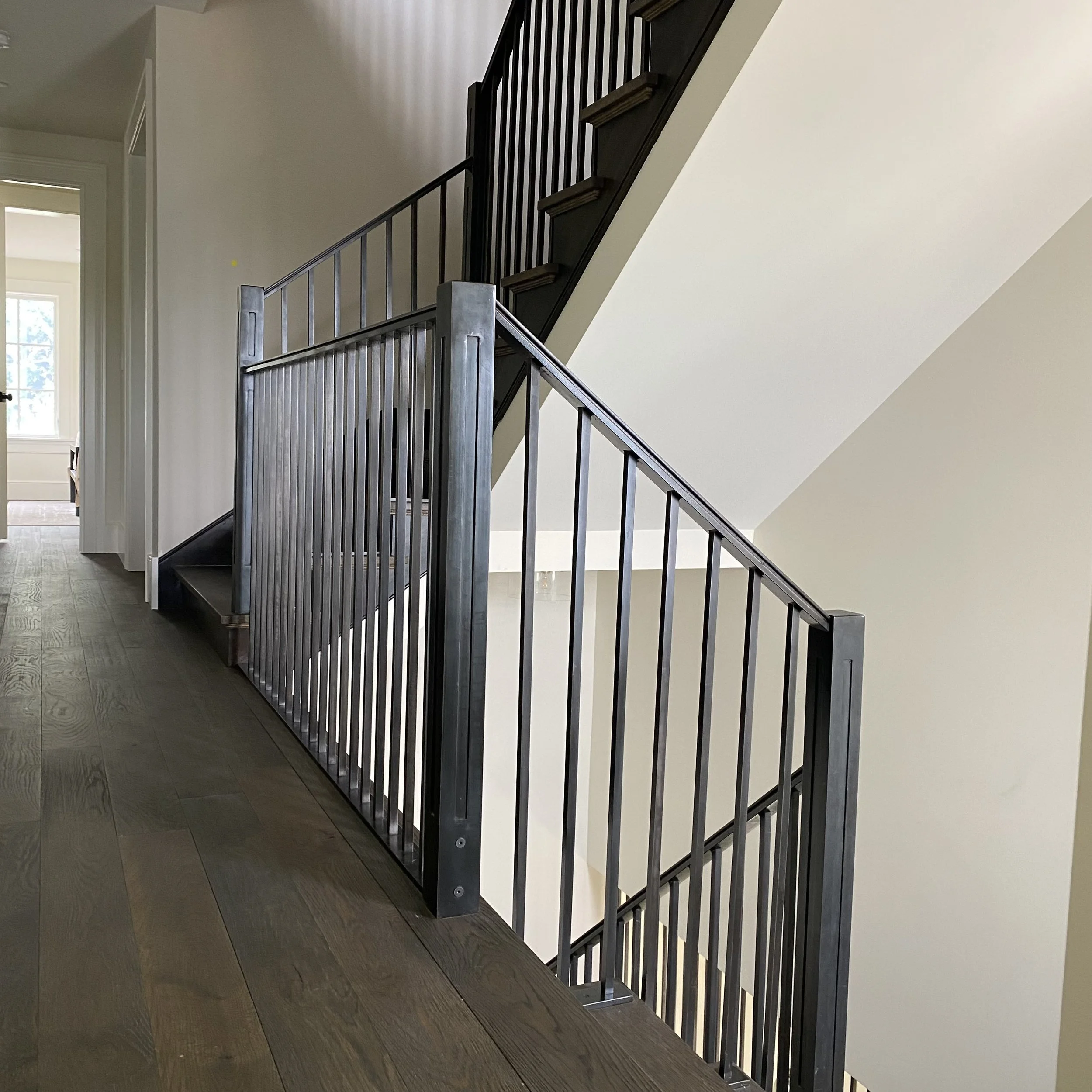 This sleek steel stair railing by Red Iron Studio features closely spaced square balusters and substantial squared newel posts for a clean, contemporary look. The blackened steel finish and streamlined design complement the dark treads and wide-plank