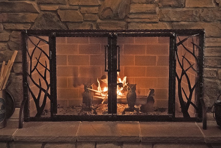 Custom hand-forged fireplace screen by Red Iron Studio, featuring organic branch-and-twig side panels with hinged doors and forged handles, glowing beautifully in front of a lit fire. This nature-inspired fire screen combines rustic artisan blacksmit