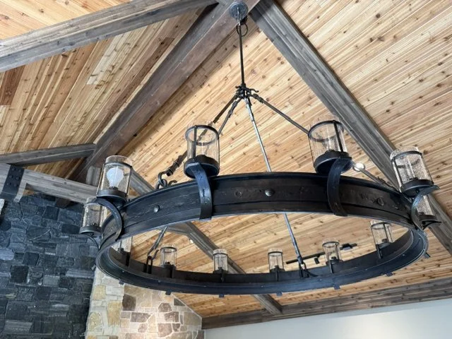 This large-scale hand-forged iron ring chandelier features a riveted circular band suspending glass hurricane candle sconces, creating a dramatic rustic-industrial lighting statement for vaulted, timber-frame ceilings. Perfect for great rooms, lodges
