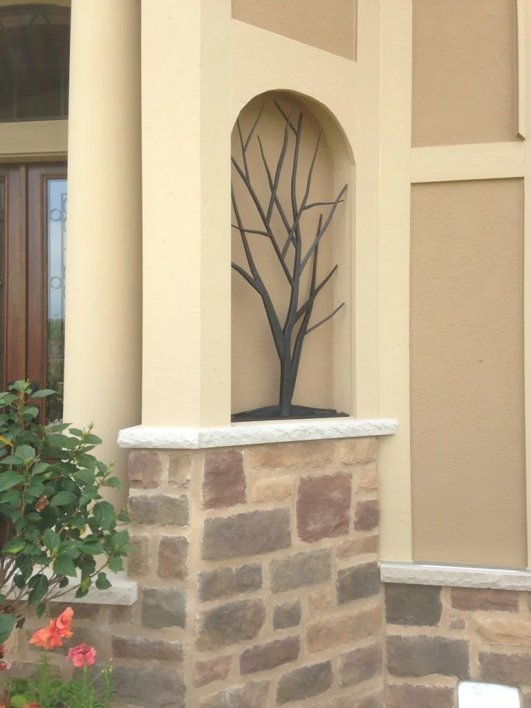This hand-forged iron tree sculpture features bare, winter-branch silhouettes set within an arched stone and stucco entryway niche, adding year-round sculptural interest to a home's exterior façade. A striking blend of nature and metalwork, this cust