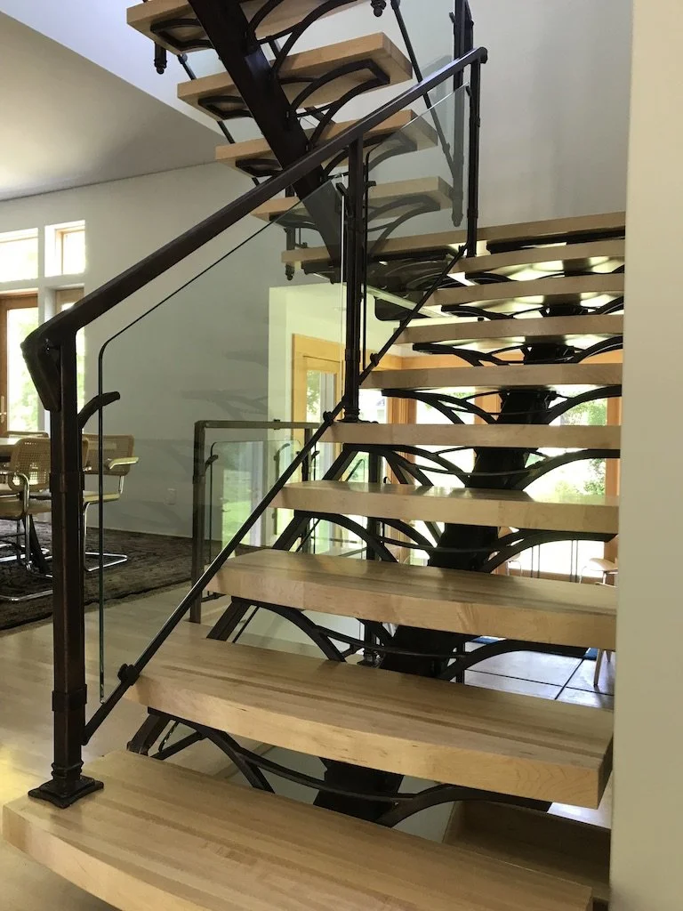 Sculptural hand-forged wrought iron staircase with a branching tree-like central support and glass panel railing by Red Iron Studio, featuring floating maple wood treads in a light-filled modern home.