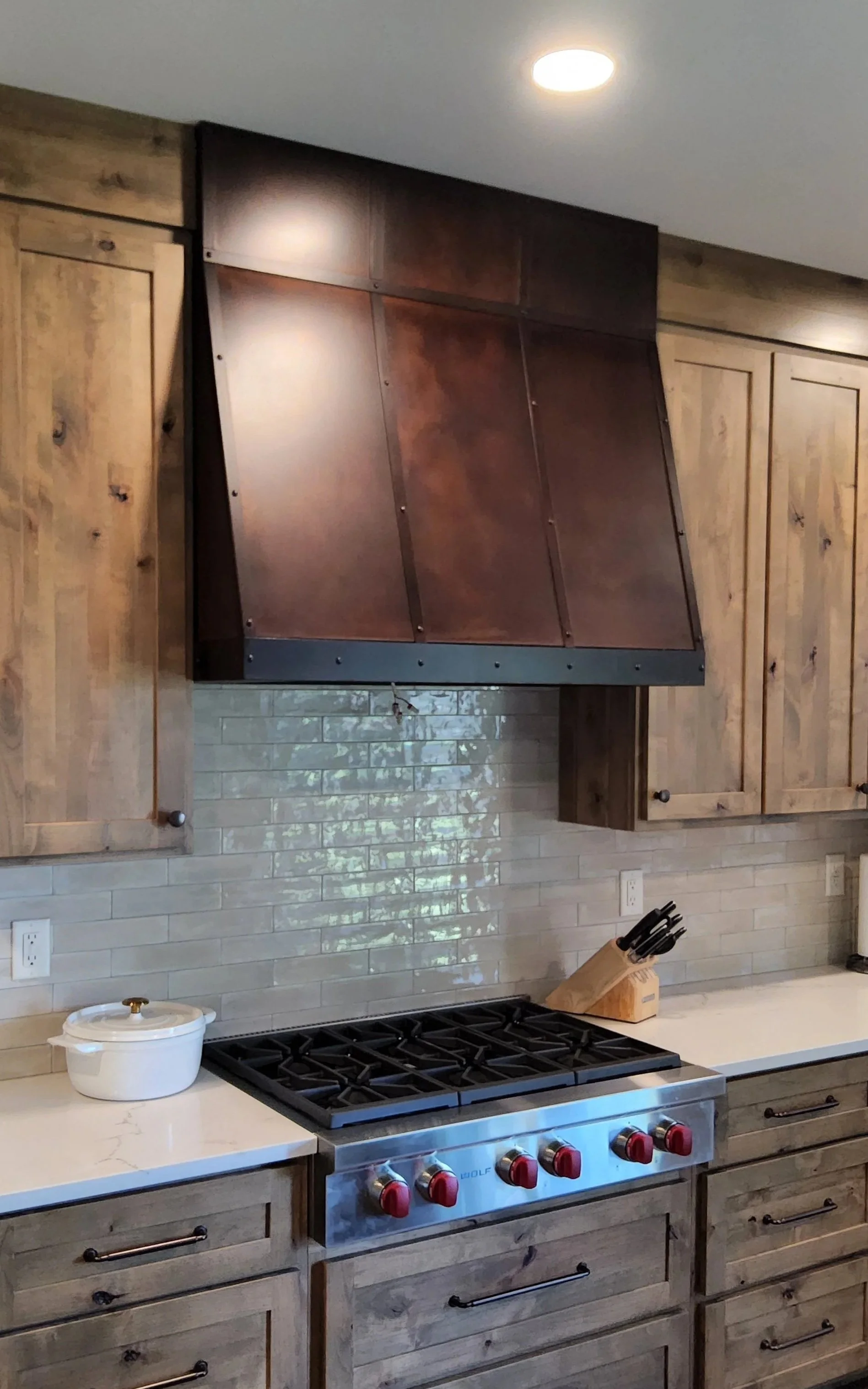 Custom copper range hood with a rich patinated finish above a Wolf gas cooktop, flanked by rustic knotty alder cabinetry and a glazed subway tile backsplash — handcrafted by Red Iron Studios.