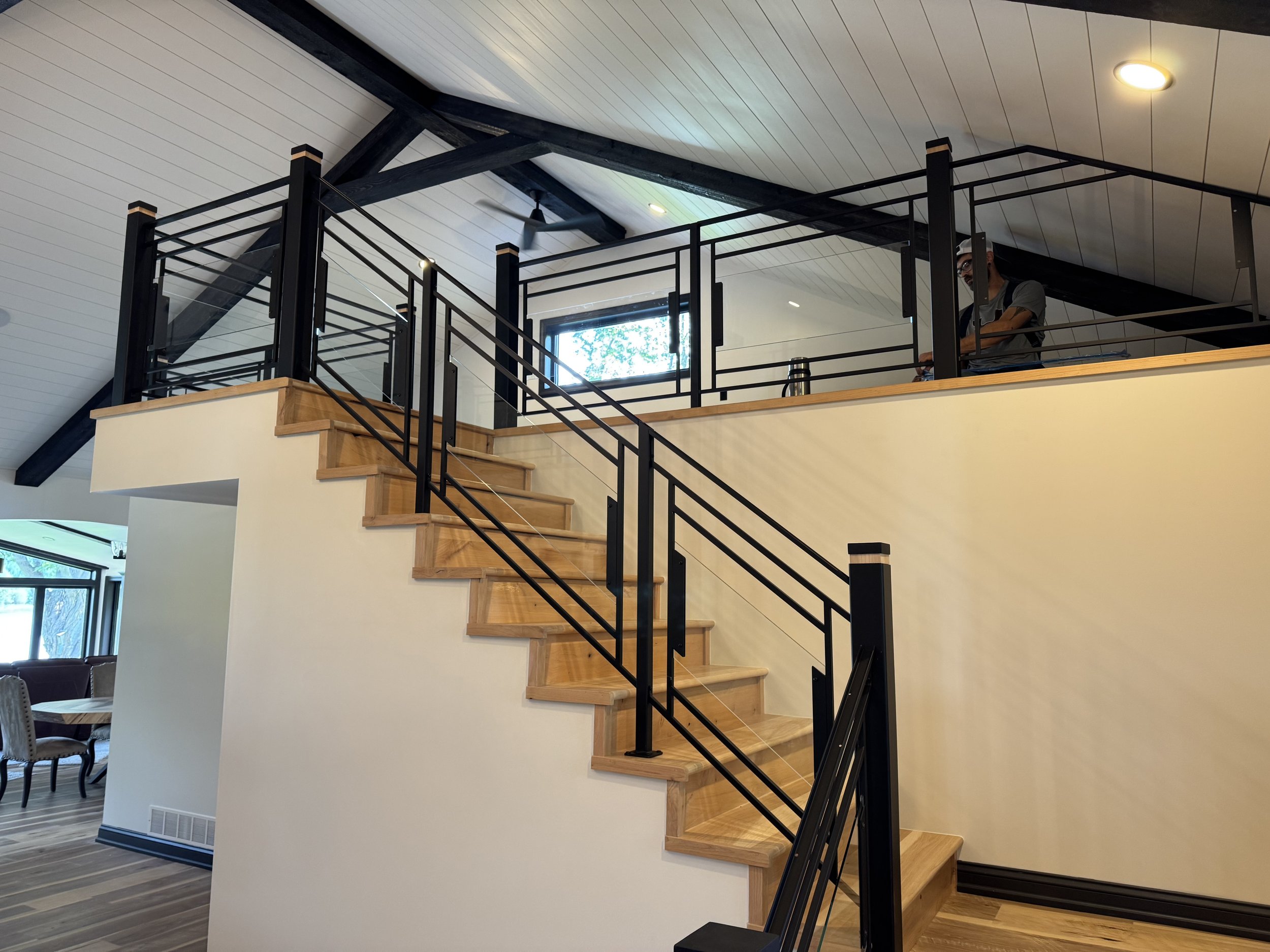 This custom black metal railing system by Red Iron Studio pairs clean horizontal lines with cable and glass infill panels, creating an open, modern look along the staircase and loft. Paired with black timber beams and warm wood treads, it brings an i