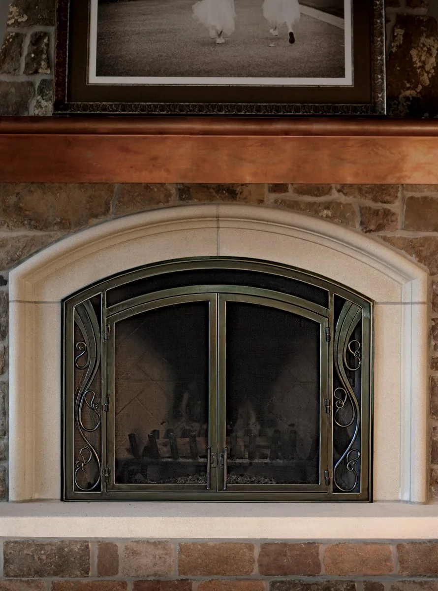 Custom arched fireplace doors by Red Iron Studio, hand-forged with elegant scrollwork side panels and a warm bronze-toned finish to complement a limestone surround. These handcrafted iron fireplace doors add graceful, traditional detail to a stacked 