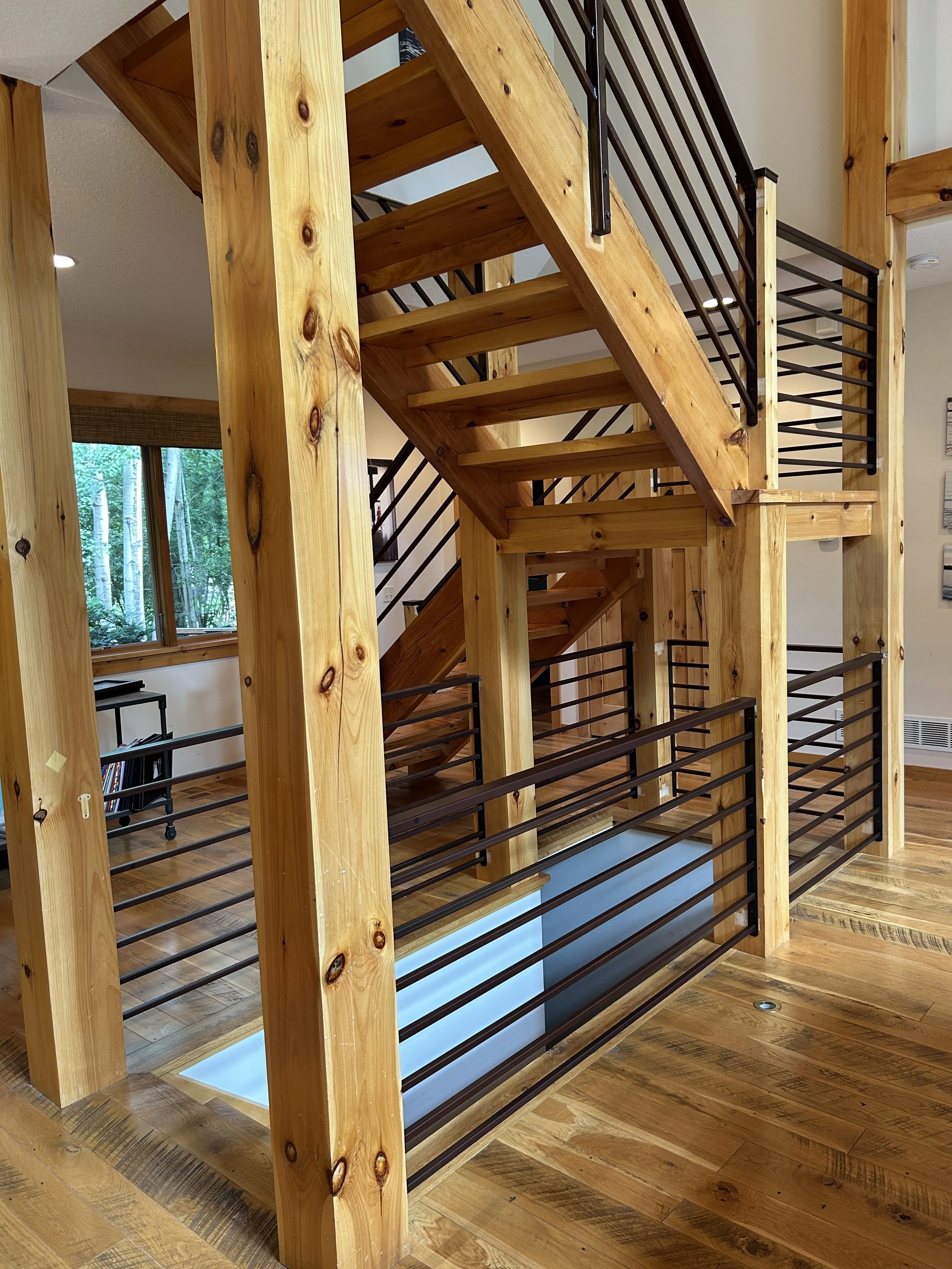 Modern black horizontal bar railing by Red Iron Studio, installed throughout a timber-frame home's open staircase and loft landings, complementing exposed wood beams and reclaimed hardwood floors.