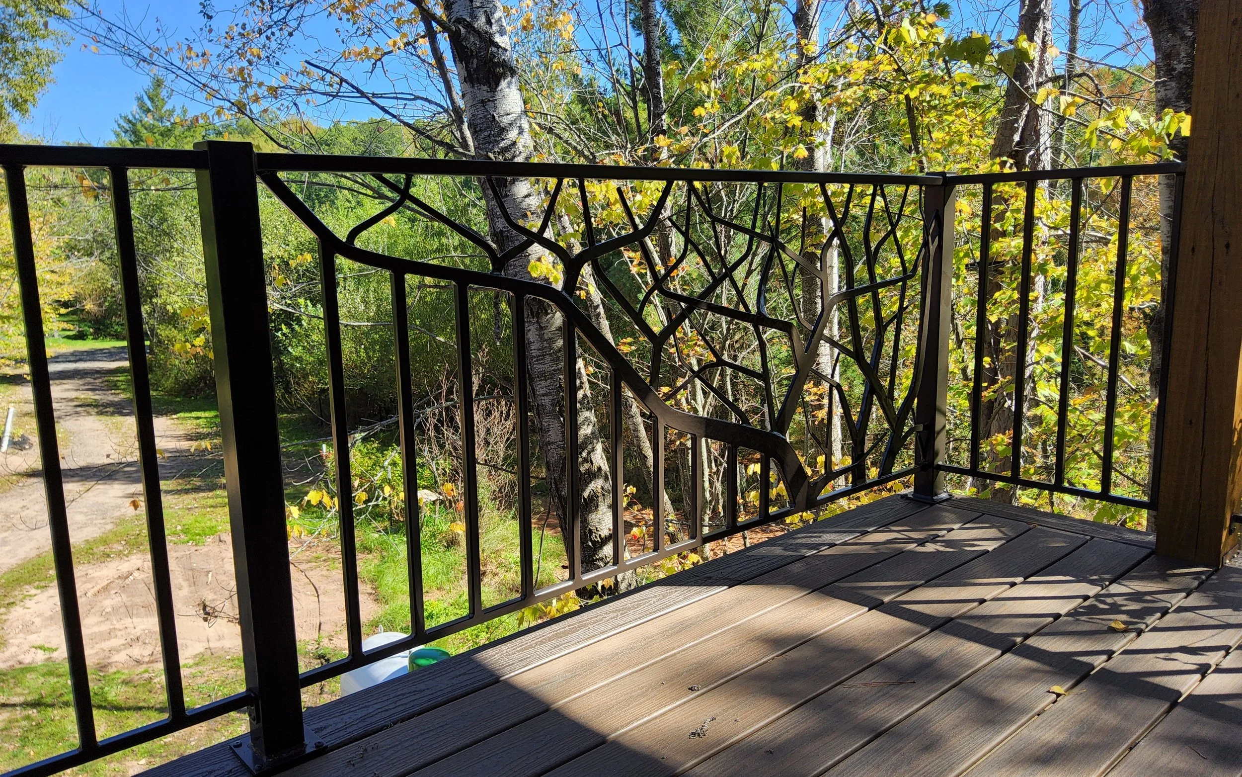 Hand-forged black wrought iron deck railing featuring a sculptural branch and twig panel design by Red Iron Studio, installed on a wooded outdoor deck overlooking birch trees and a gravel path.