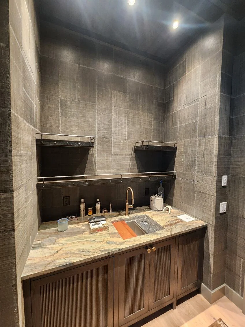 This custom blackened steel shelving system features sleek floating niches with riveted rail detailing, built into a wet bar nook for a sophisticated, industrial-modern display of glassware and barware. Paired with natural stone countertops, this cus