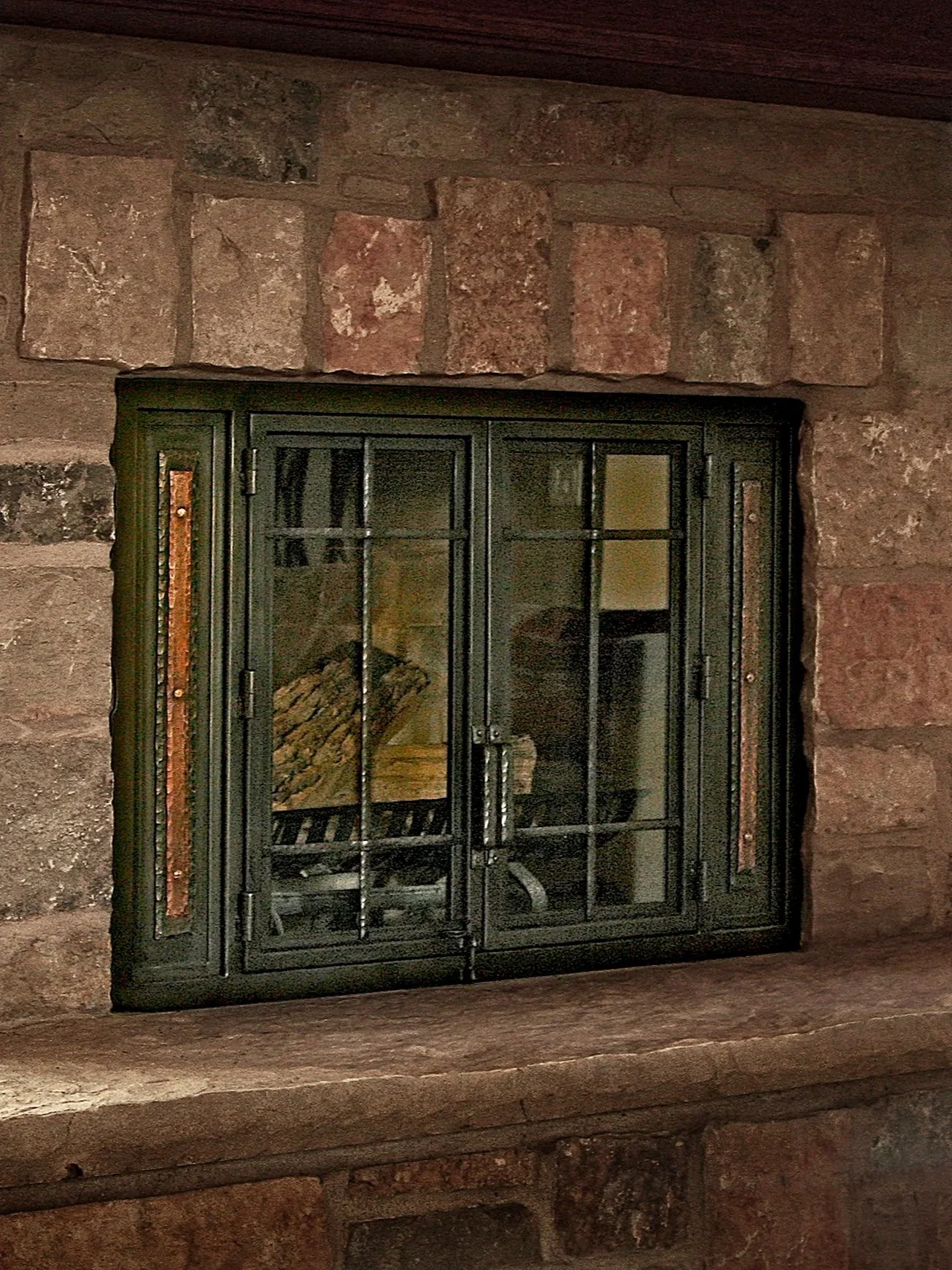 Custom black iron fireplace doors by Red Iron Studio with divided glass windows and hammered copper side panels, set into a rustic stone fireplace.