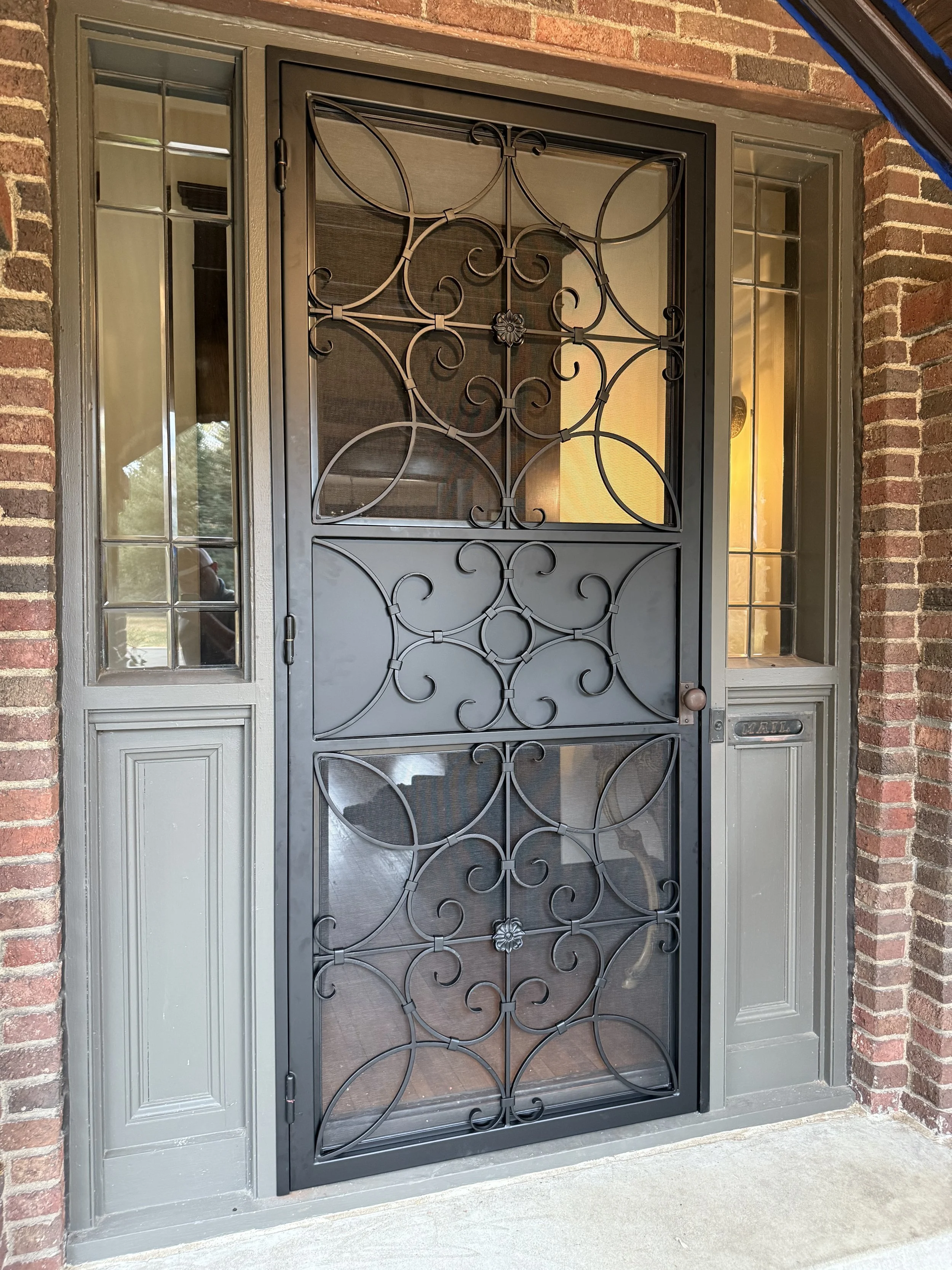 This custom wrought iron security screen door features an elegant circular and scroll pattern with a decorative floral medallion, combining protective functionality with timeless Old World charm. Perfect for traditional brick homes, this handcrafted 