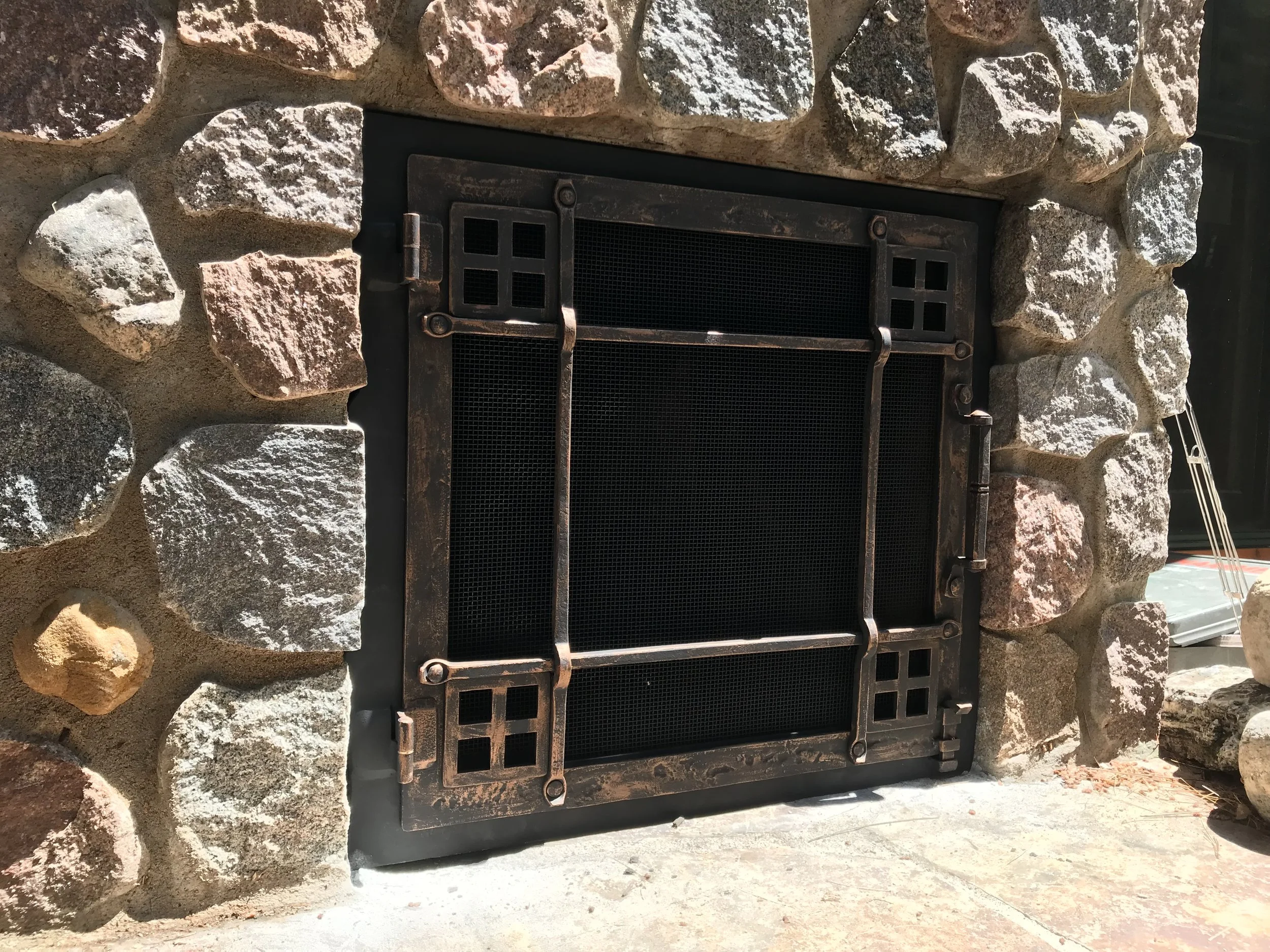 Custom Craftsman-style fireplace screen by Red Iron Studio, hand-forged with grid mullion corners and a hand-hammered copper-toned finish, set into a rugged fieldstone outdoor fireplace. This handcrafted fire screen brings warm artisan detail and tra