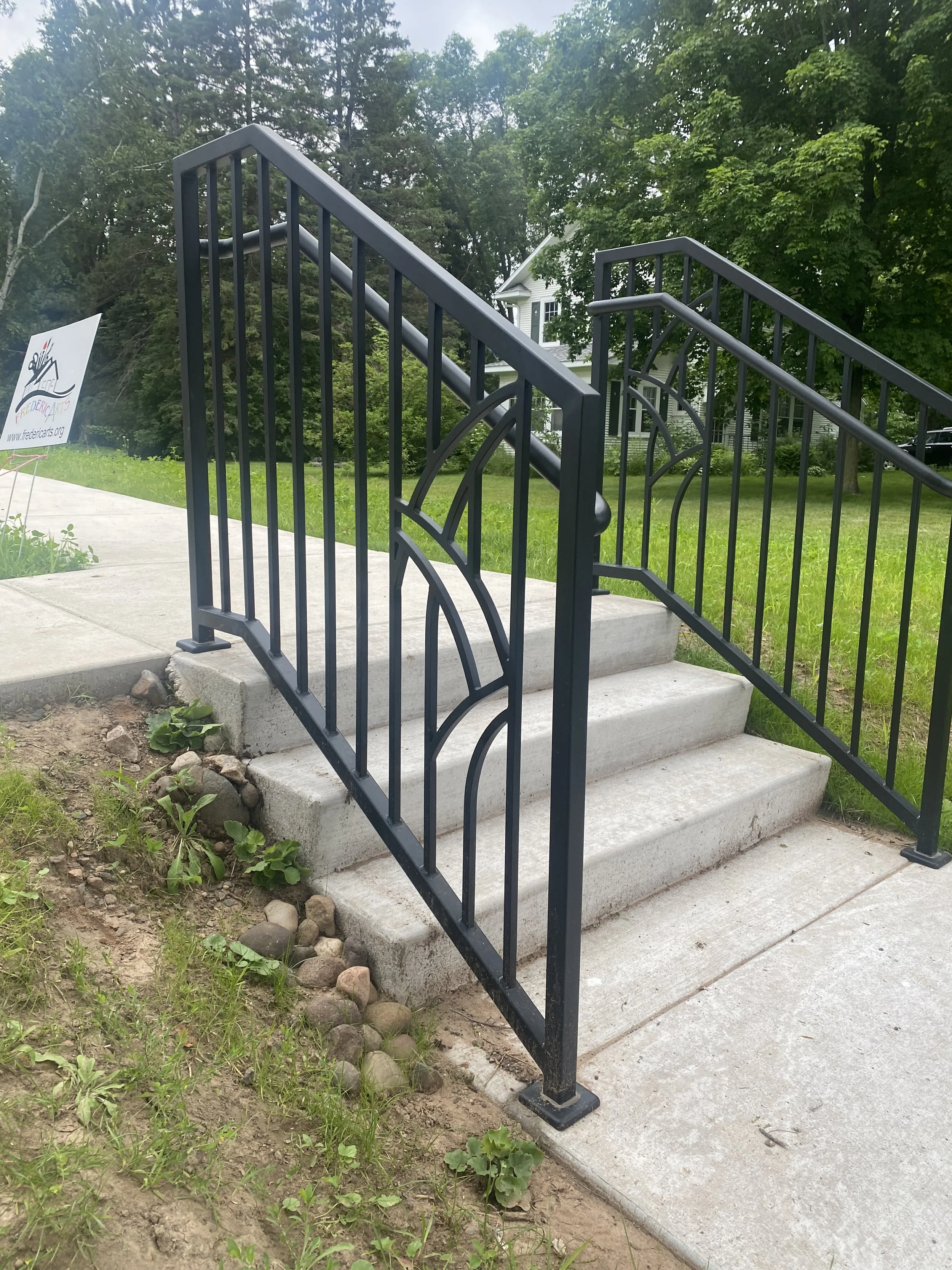 Custom black steel outdoor stair railing featuring a sunburst arch panel design, fabricated by Red Iron Studio for a public sidewalk entrance. This durable, decorative metal handrail combines code-compliant safety with artistic flair, adding a welcom