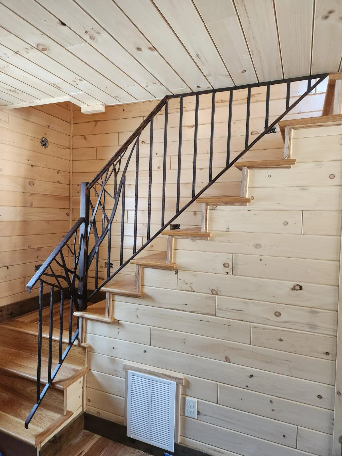 Hand-forged wrought iron stair railing featuring a sculptural branch newel post design by Red Iron Studio, paired with straight iron pickets and wood treads in a rustic knotty pine log cabin staircase.