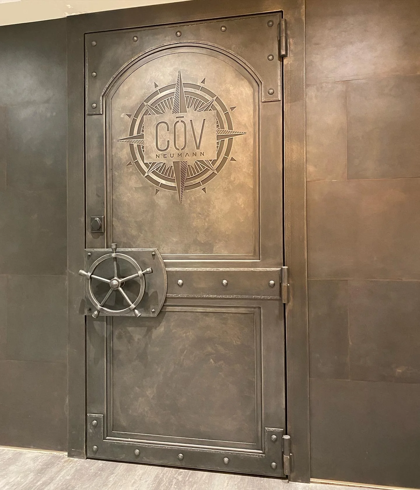 Custom Hand-Forged Steel Vault Door with Engraved Compass Logo - This custom steel vault door features a riveted, industrial design with a hand-cut compass rose logo and functional ship's wheel locking mechanism, creating a striking nautical-themed e