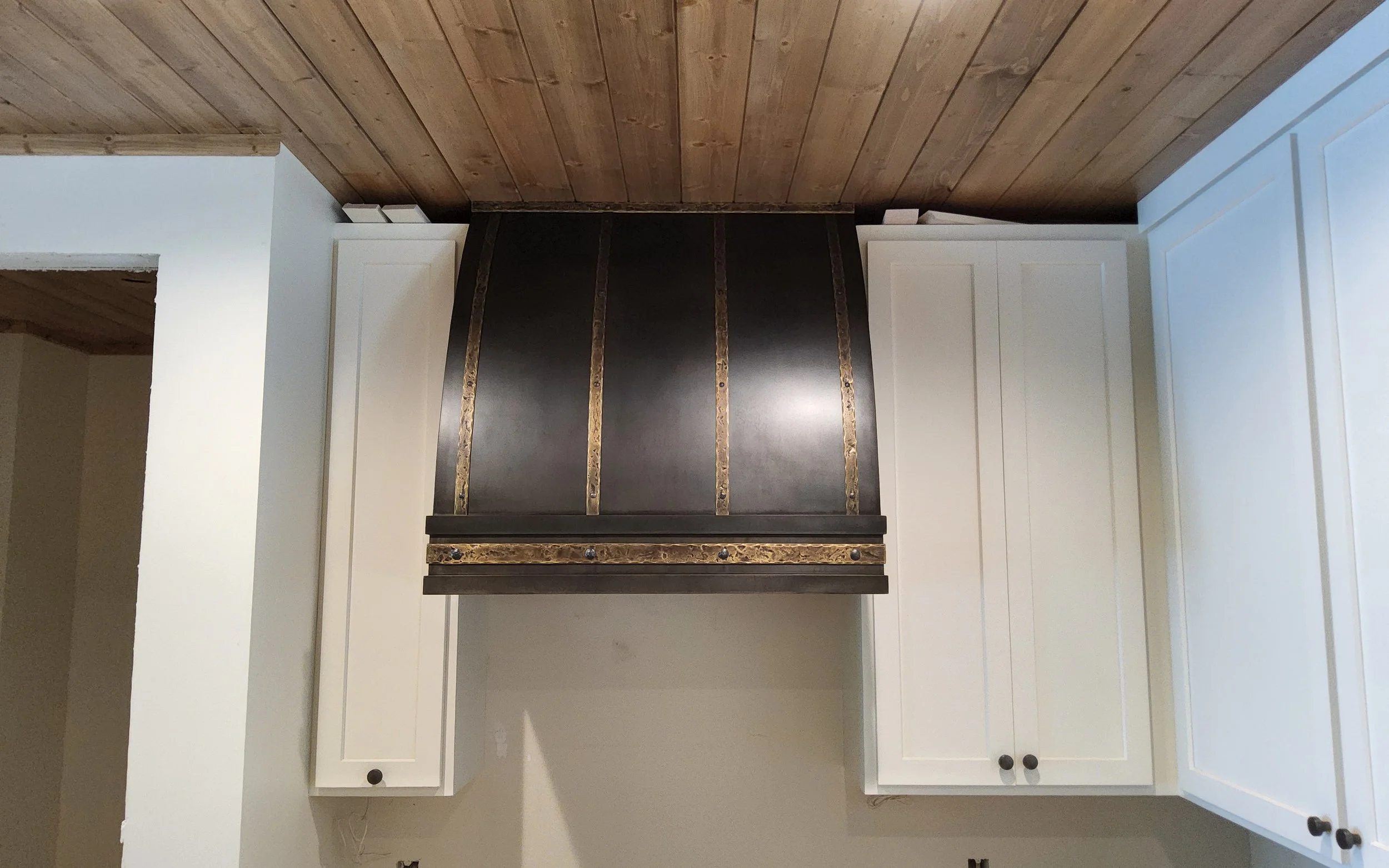 Custom Blackened Steel Range Hood with Hammered Bronze Strapping - This gracefully curved range hood pairs a deep blackened steel finish with hand-hammered bronze strapping and rivet detail, creating a refined yet rustic centerpiece beneath a tongue-