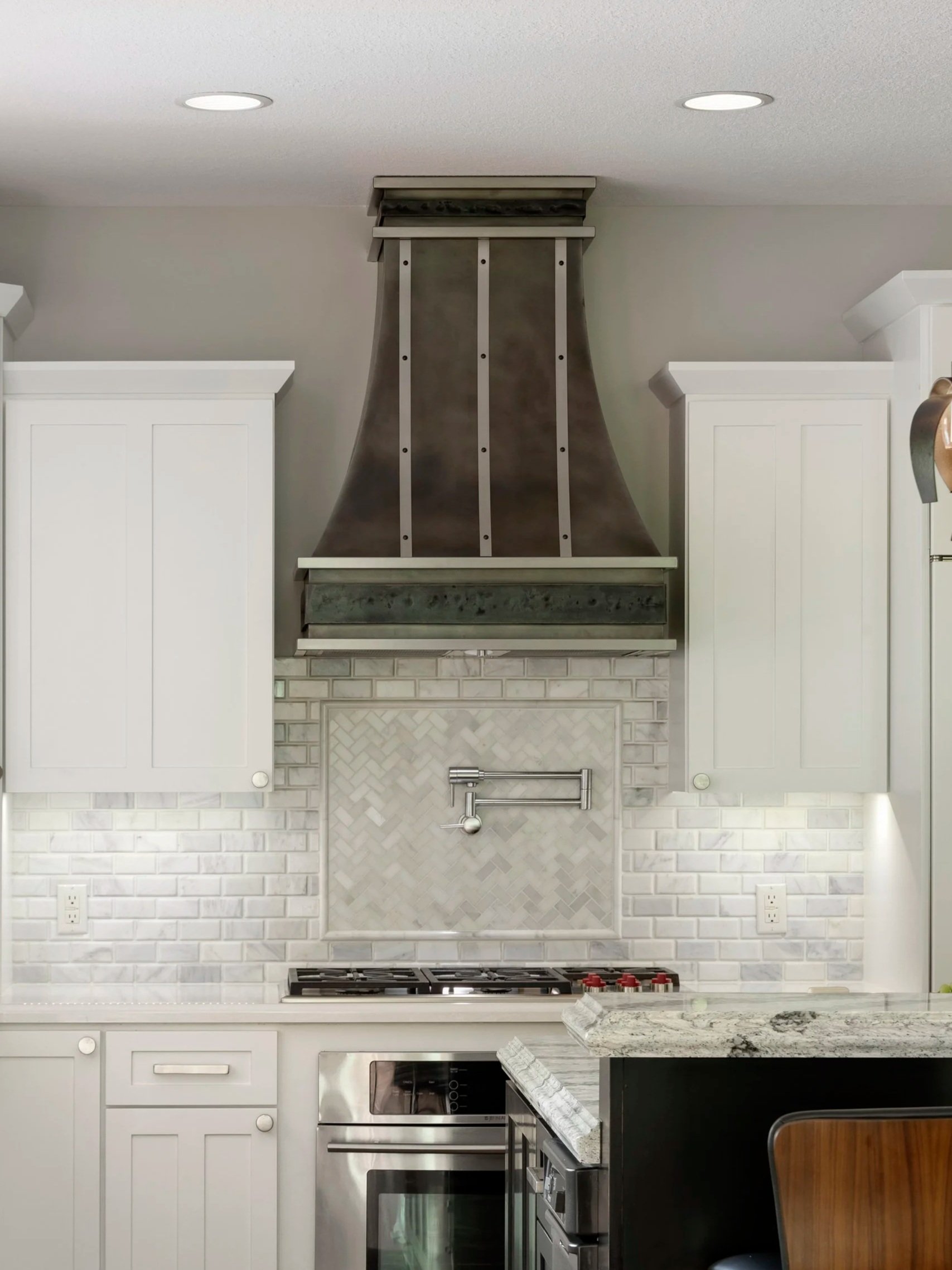 Curved stainless steel range hood with vertical rivets over a herringbone marble tile backsplash and gas cooktop.