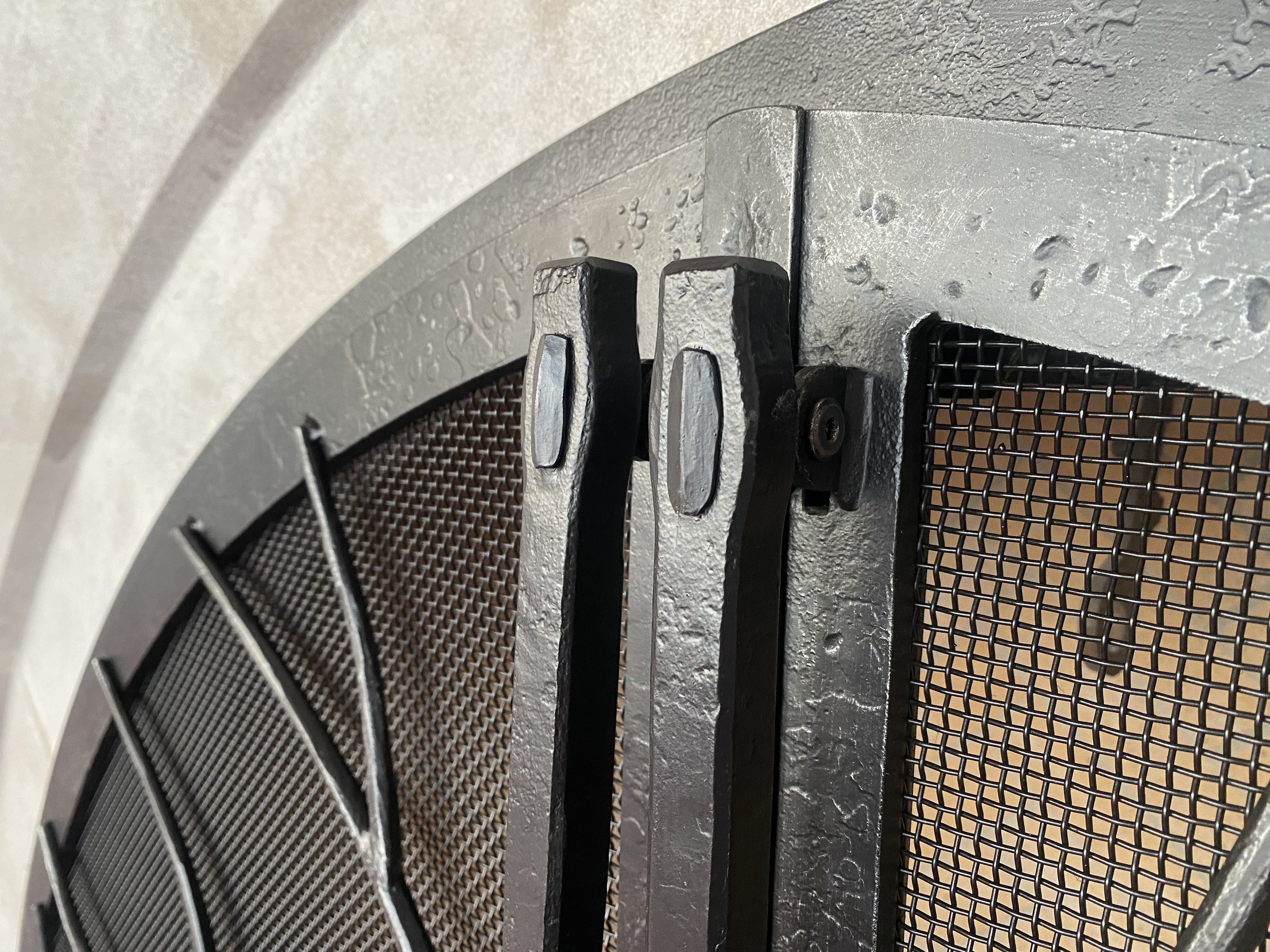 Close-up detail of a custom hand-forged fireplace door handle by Red Iron Studio, showing hammer-textured iron and a hand-riveted joint set against curved mesh panels. This detail image highlights the meticulous artisan blacksmithing behind Red Iron 