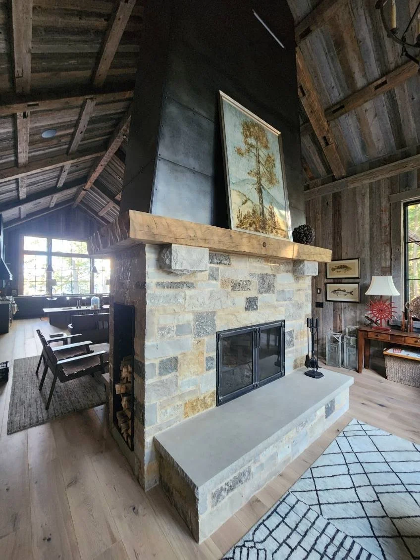 Custom blackened steel chimney surround by Red Iron Studio, hand-fabricated with a tall riveted metal chase above a stacked stone fireplace, paired with clean-lined fireplace doors and a matching log bin. This custom chimney and fireplace door combin
