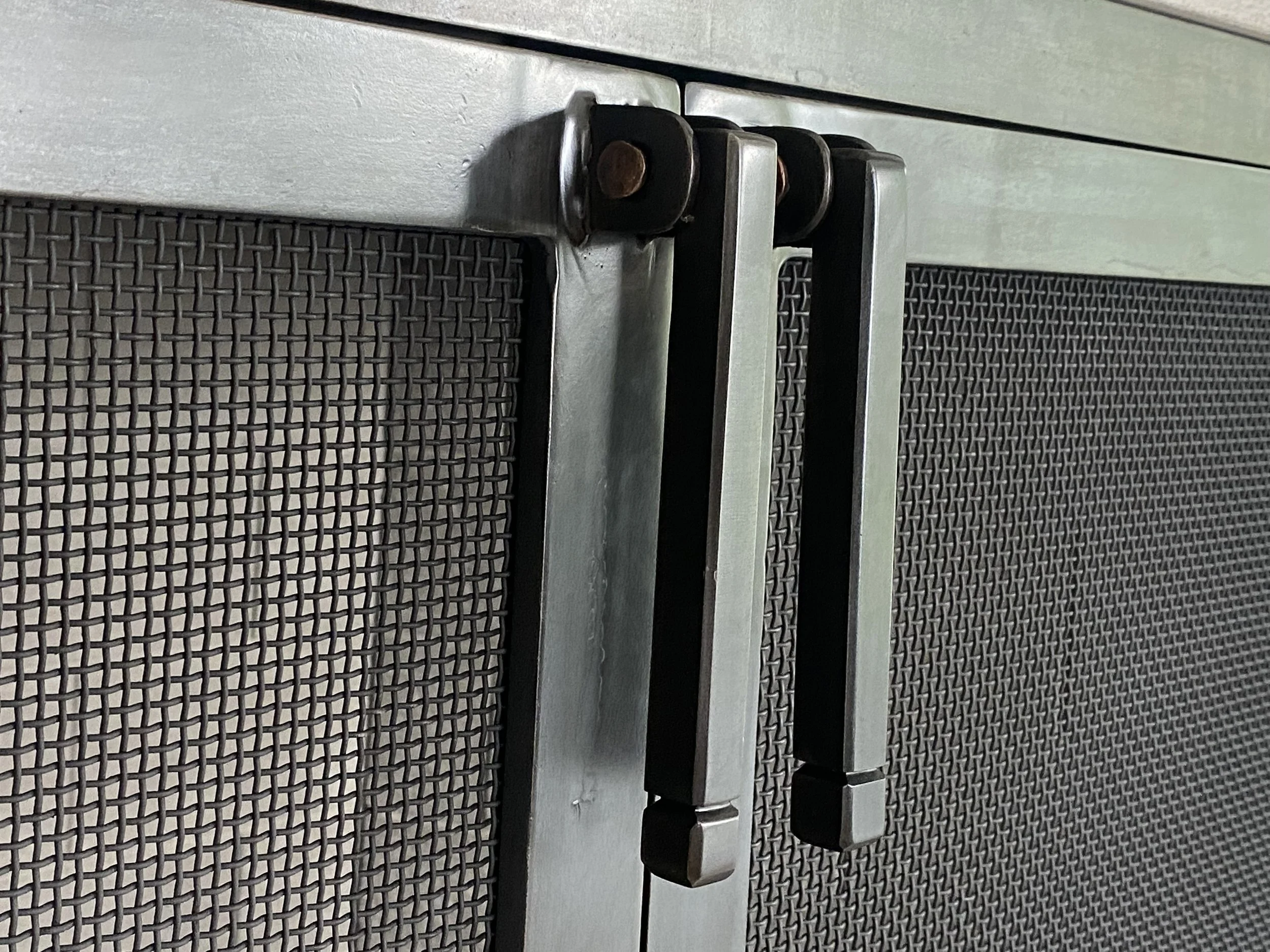 Close-up detail of custom hand-forged fireplace door handles by Red Iron Studio, showcasing precision blacksmithing with copper rivet accents and a clean, squared iron profile against fine mesh screen panels. This detail shot highlights the fine craf