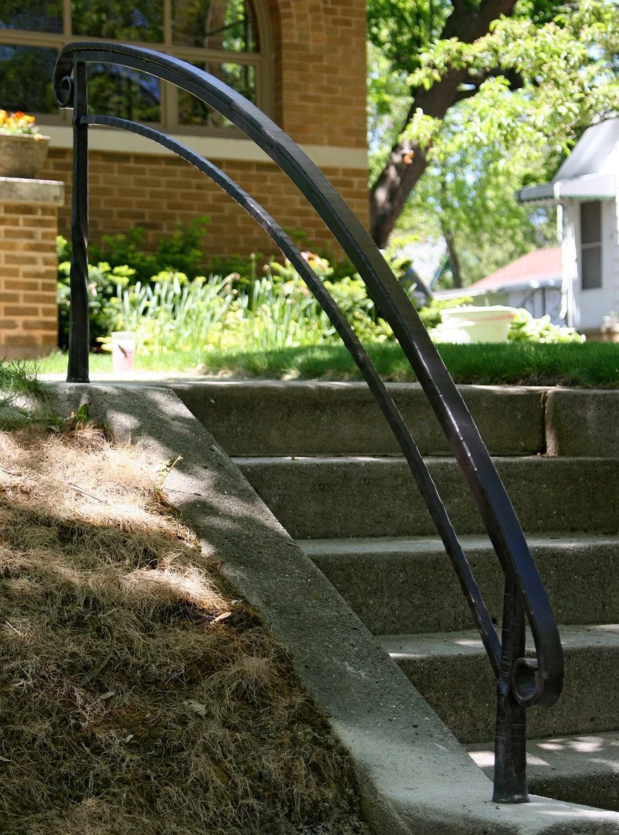 Custom hand-forged wrought iron handrail with a sweeping arched design and scrolled ends by Red Iron Studio, installed on a brick building's concrete entrance steps.
