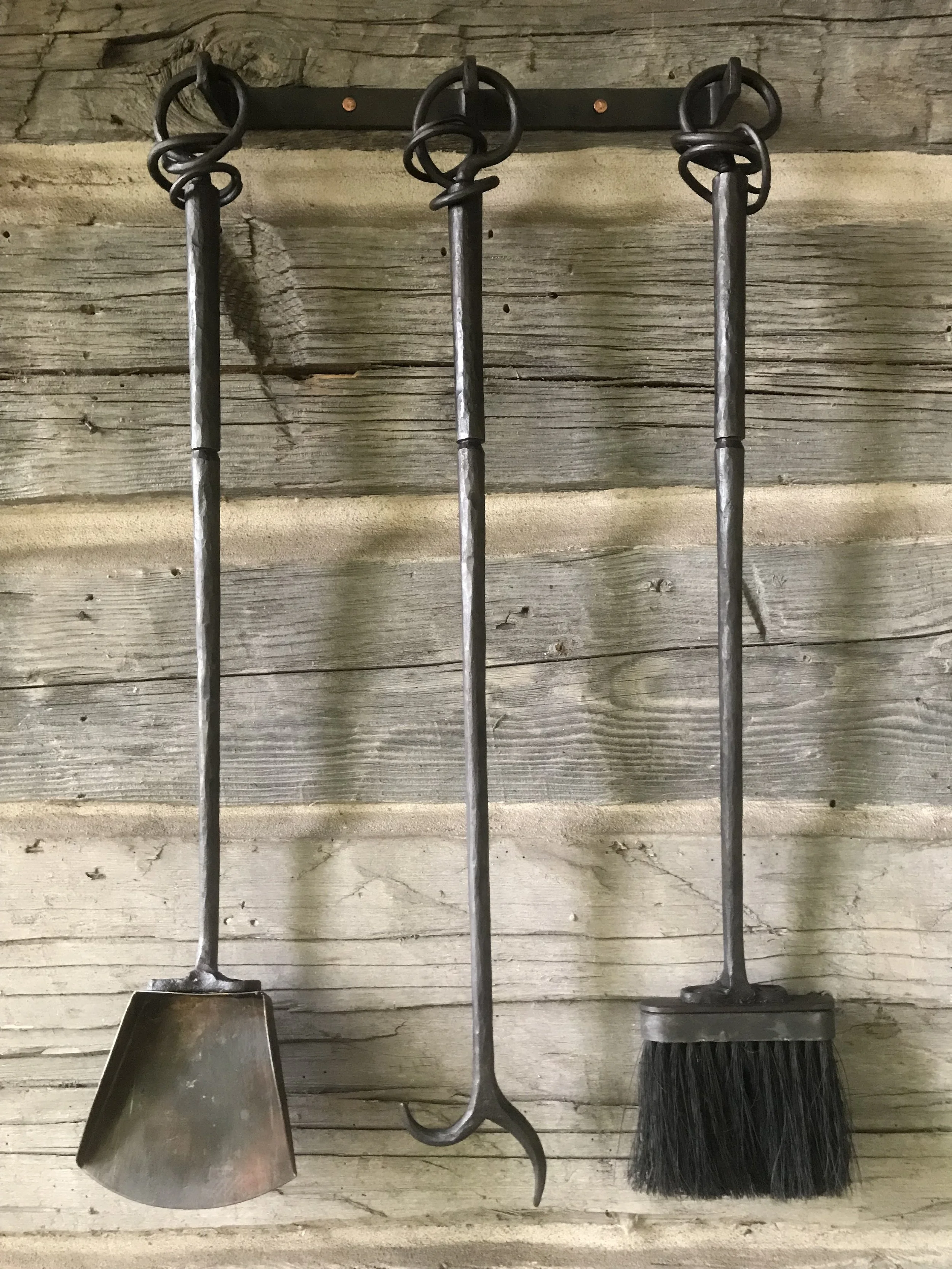 Hand-forged fireplace tool set by Red Iron Studio, featuring a shovel, poker, and brush with distinctive hand-twisted iron ring handles and a matching wall-mounted hook rail. This custom fireplace accessory set showcases traditional blacksmithing cra