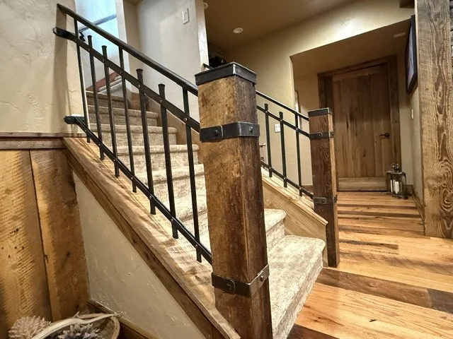 Custom wrought iron stair railing with hand-forged balusters and rustic wood newel posts wrapped in iron strapping by Red Iron Studio, installed on a carpeted staircase in a timber-accented home.
