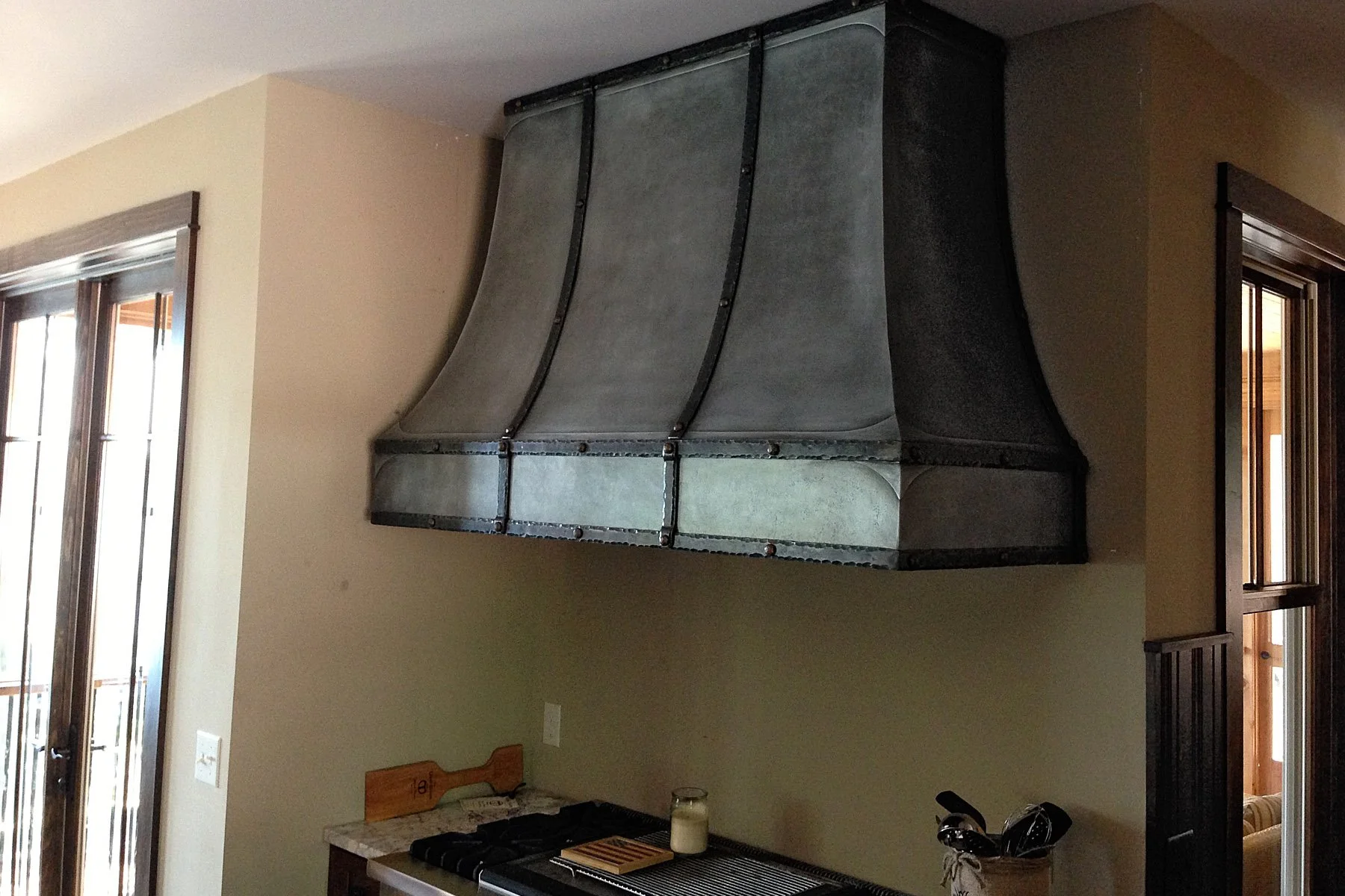 Custom zinc-finish range hood with vertical iron strapping and hand-riveted seams, angled above a cooktop between timber-framed windows handcrafted by Red Iron Studios.