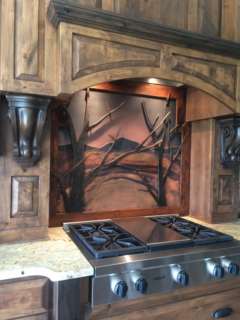 This custom metal stove backsplash features a hand-forged mountain landscape scene with dimensional iron tree branches rising in relief against a rich patinated copper panel, creating a dramatic focal point for the kitchen range. A true work of funct