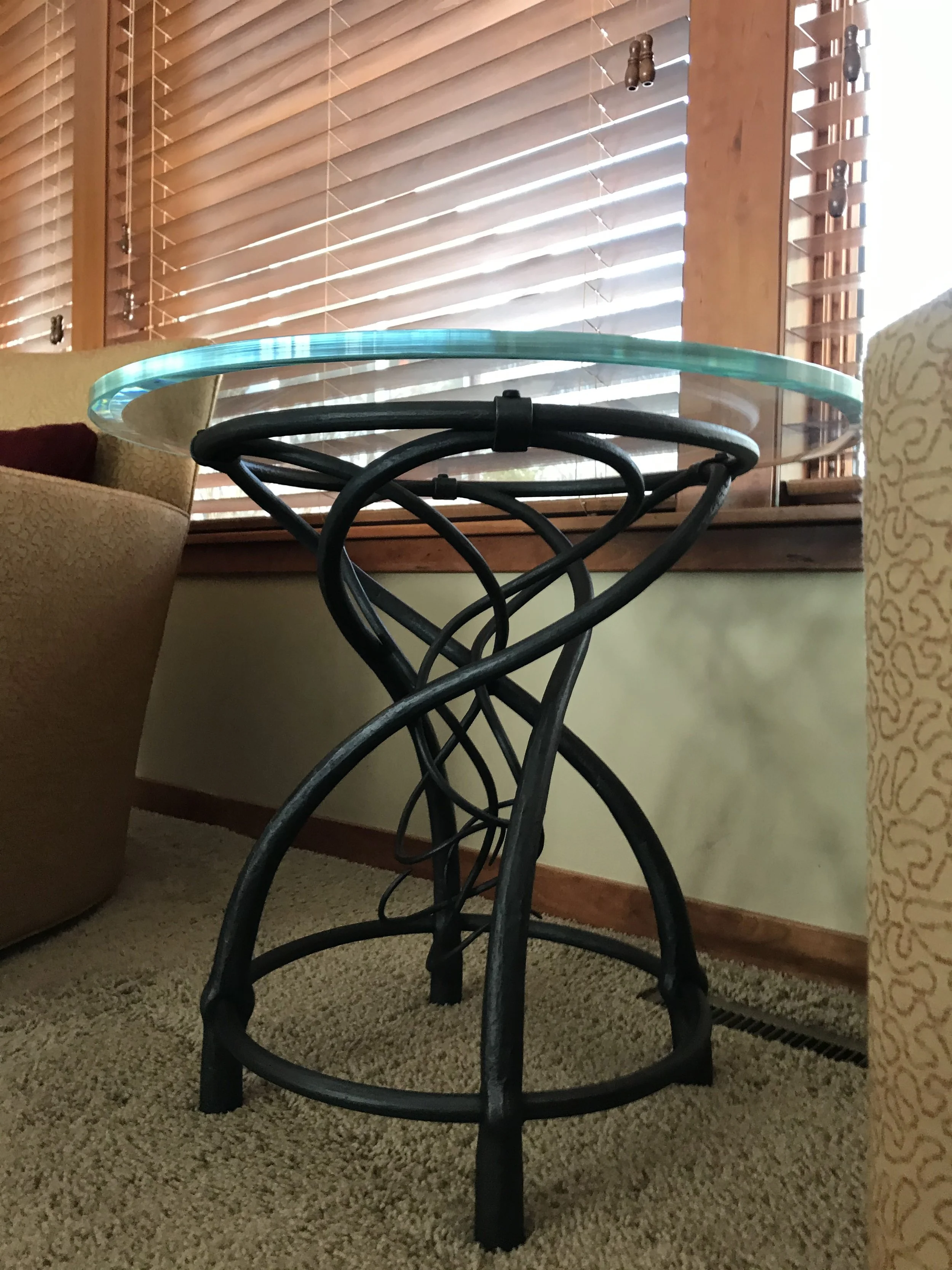 Hand-Forged Iron Side Table with Glass Top - This artisan-crafted iron side table features an intricately woven, hourglass-shaped base of hand-forged steel topped with a round tempered glass surface, blending sculptural metalwork with modern function