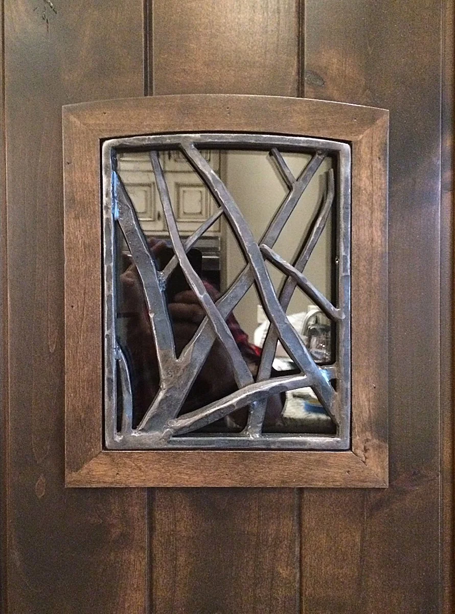This hand-forged iron window grille features an abstract, branch-like lattice pattern set within a wood-framed door window, adding artistic security and natural texture to any entryway. A striking blend of form and function, this custom iron grille s