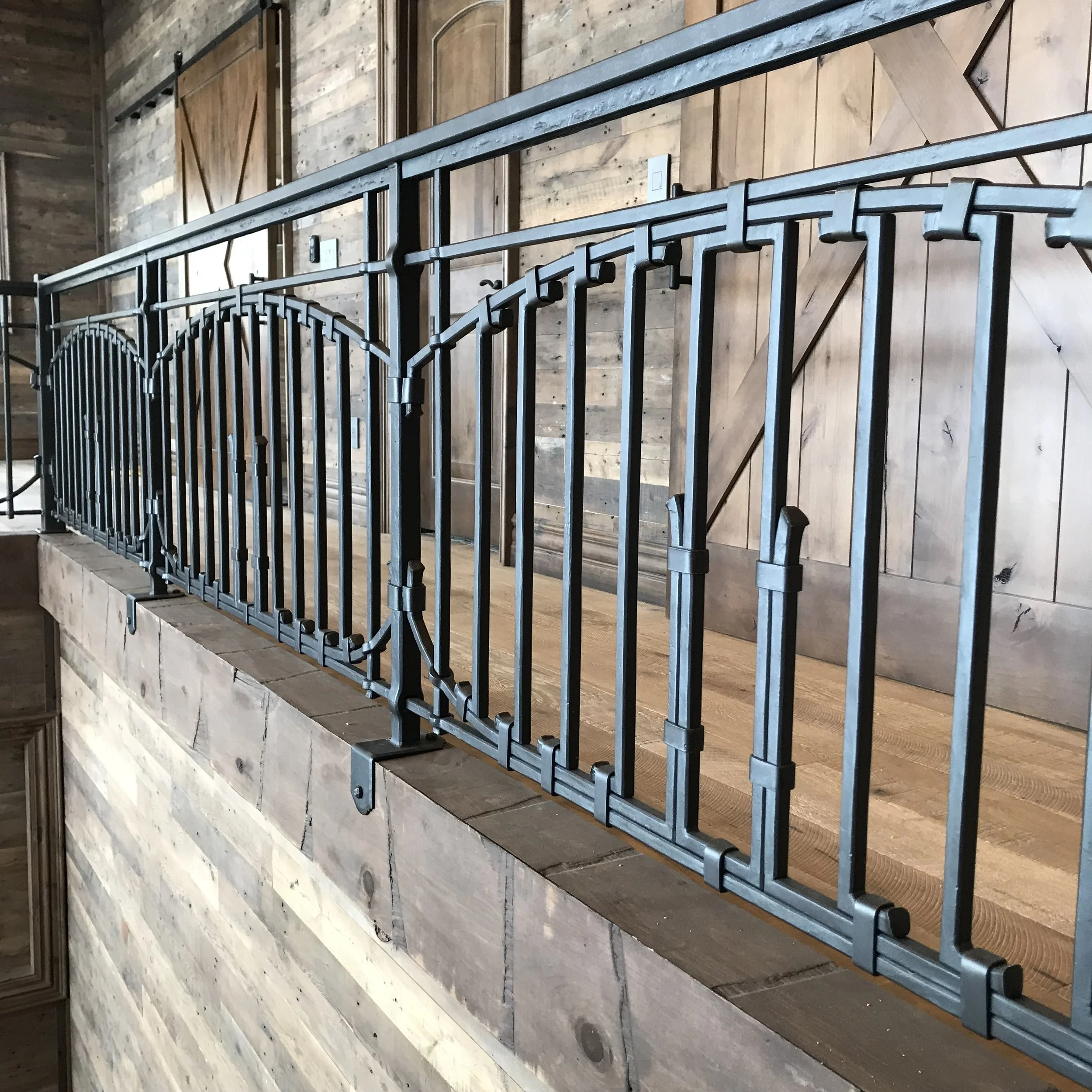 Hand-forged wrought iron railing with arched paneling and a hammered, textured finish, custom-built by Red Iron Studio for a rustic barnwood-clad loft. This artisan blacksmith railing pairs beautifully with reclaimed wood walls and wide-plank floorin