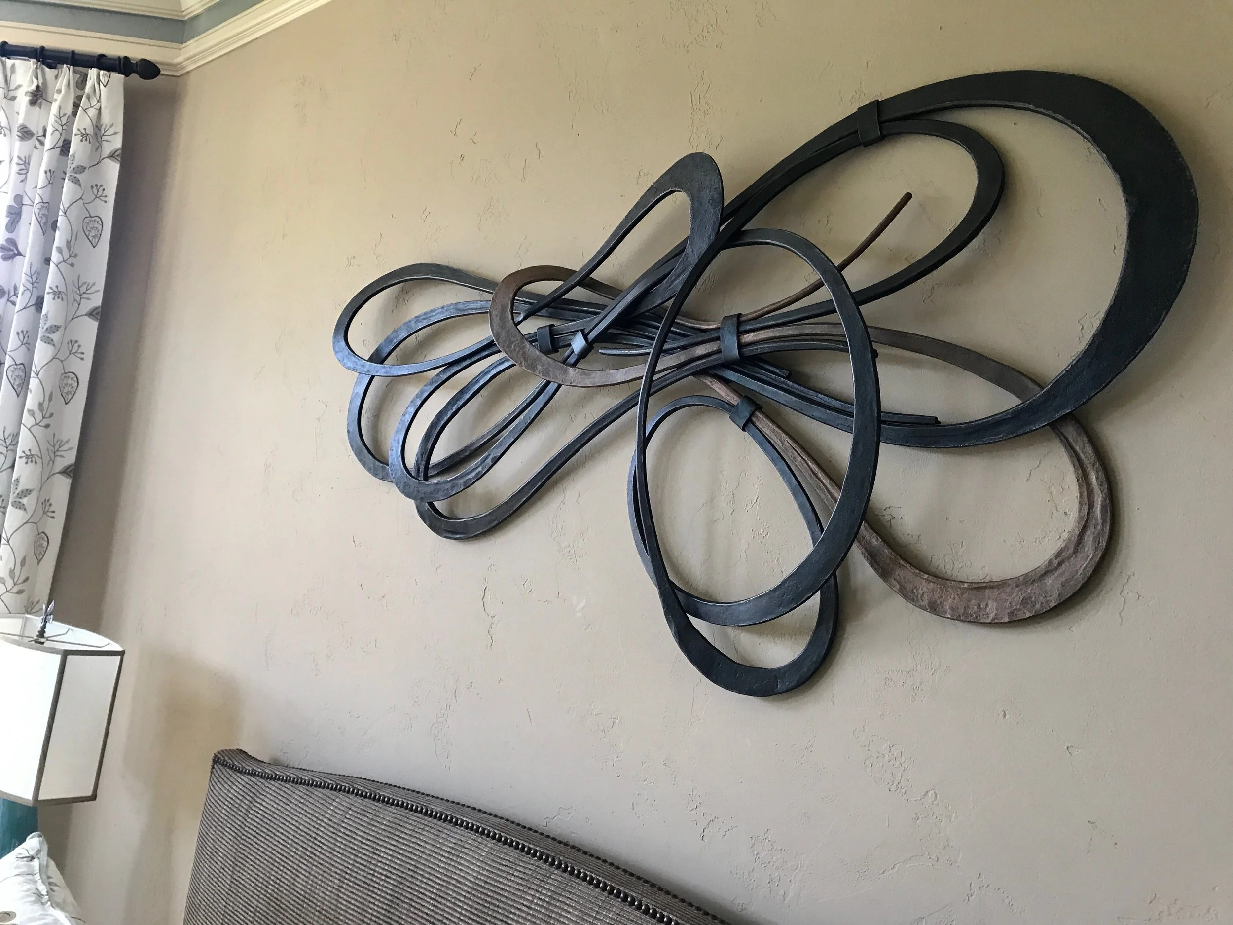 This large-scale abstract metal wall sculpture features flowing, interlacing steel ribbons in blackened and natural finishes, creating a dynamic sense of movement and depth above any sofa or focal wall. A striking statement piece for contemporary int