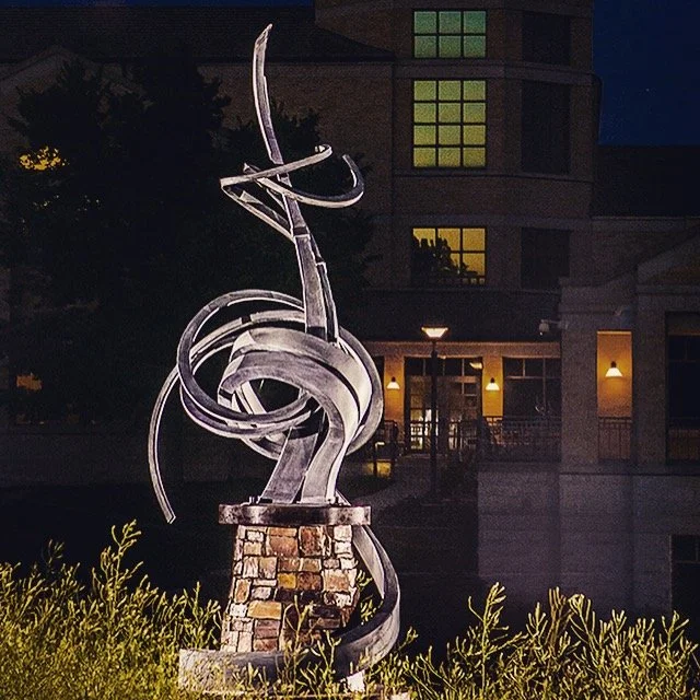 This monumental abstract steel sculpture features sweeping, ribbon-like forged forms atop a natural stone pedestal, creating a striking public art installation for civic plazas, campuses, or corporate landscapes.