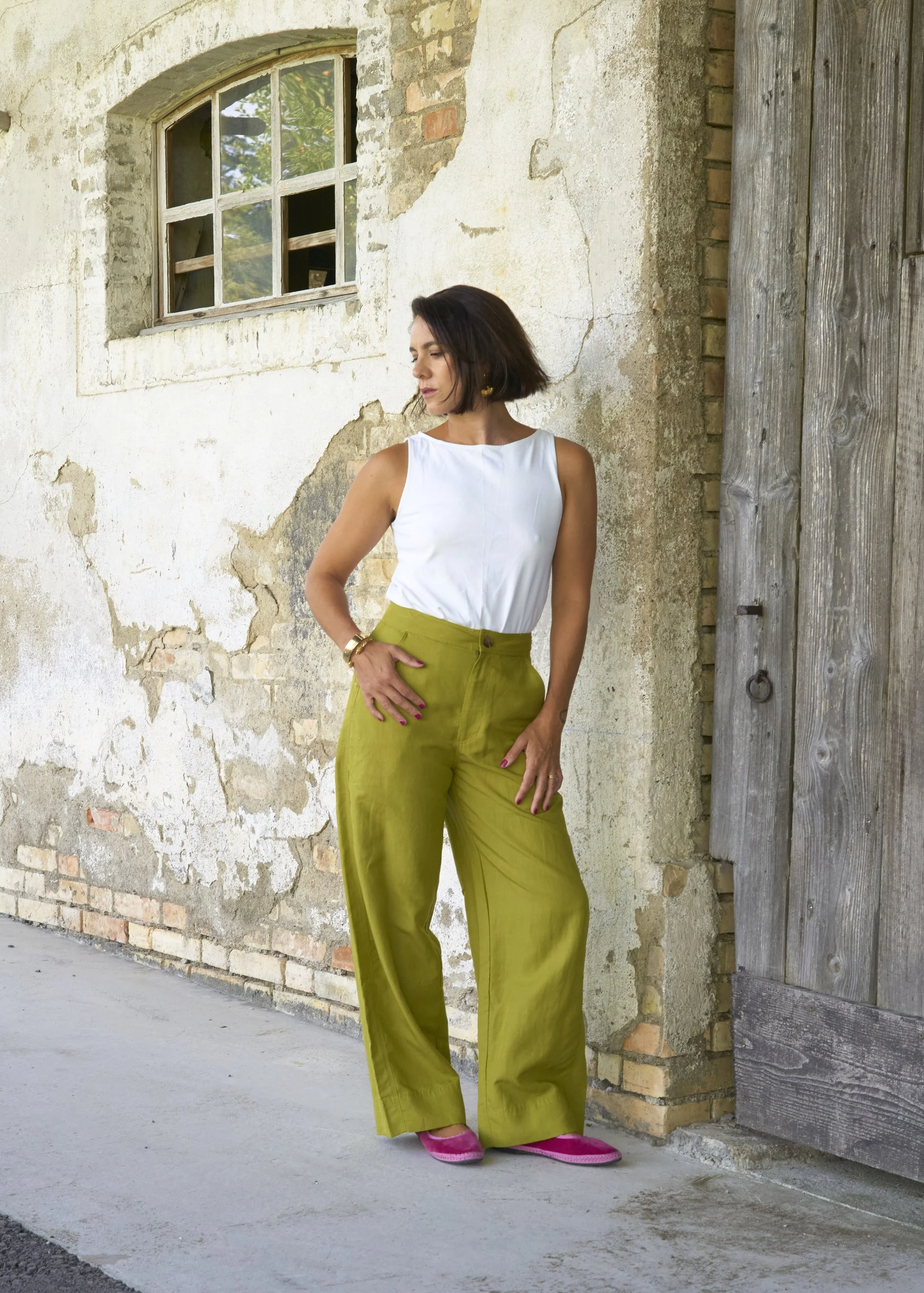 A woman wearing a white sleeveless top and bright yellow-green wide-leg pants, standing against an aged, textured wall with a window and a wooden door.