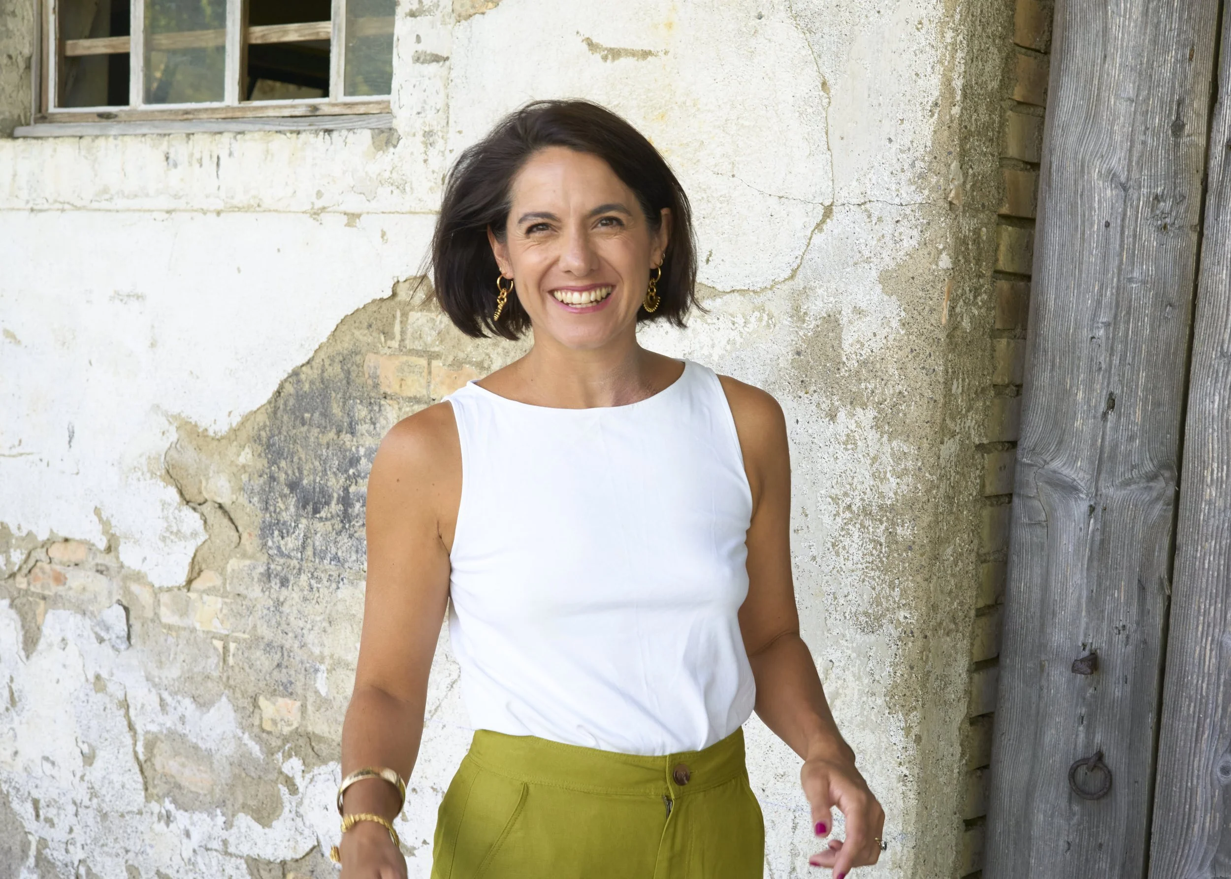 A woman smiling outdoors standing in front of a weathered brick wall and wooden fence.