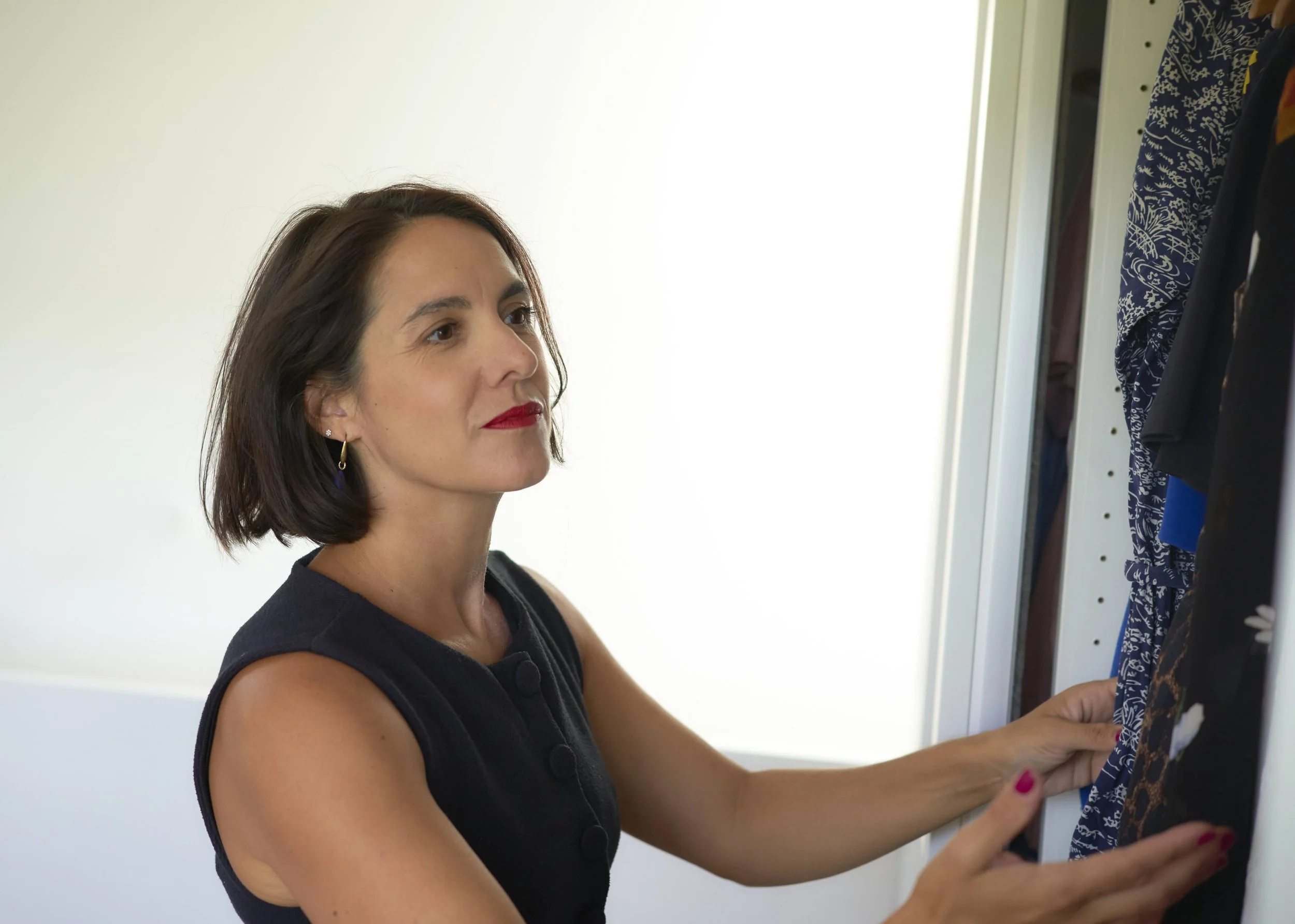 A woman with shoulder-length dark hair, red lipstick, and earrings is looking at clothing items hanging on a closet or wardrobe. She is wearing a sleeveless black top and is touching or holding a piece of clothing.