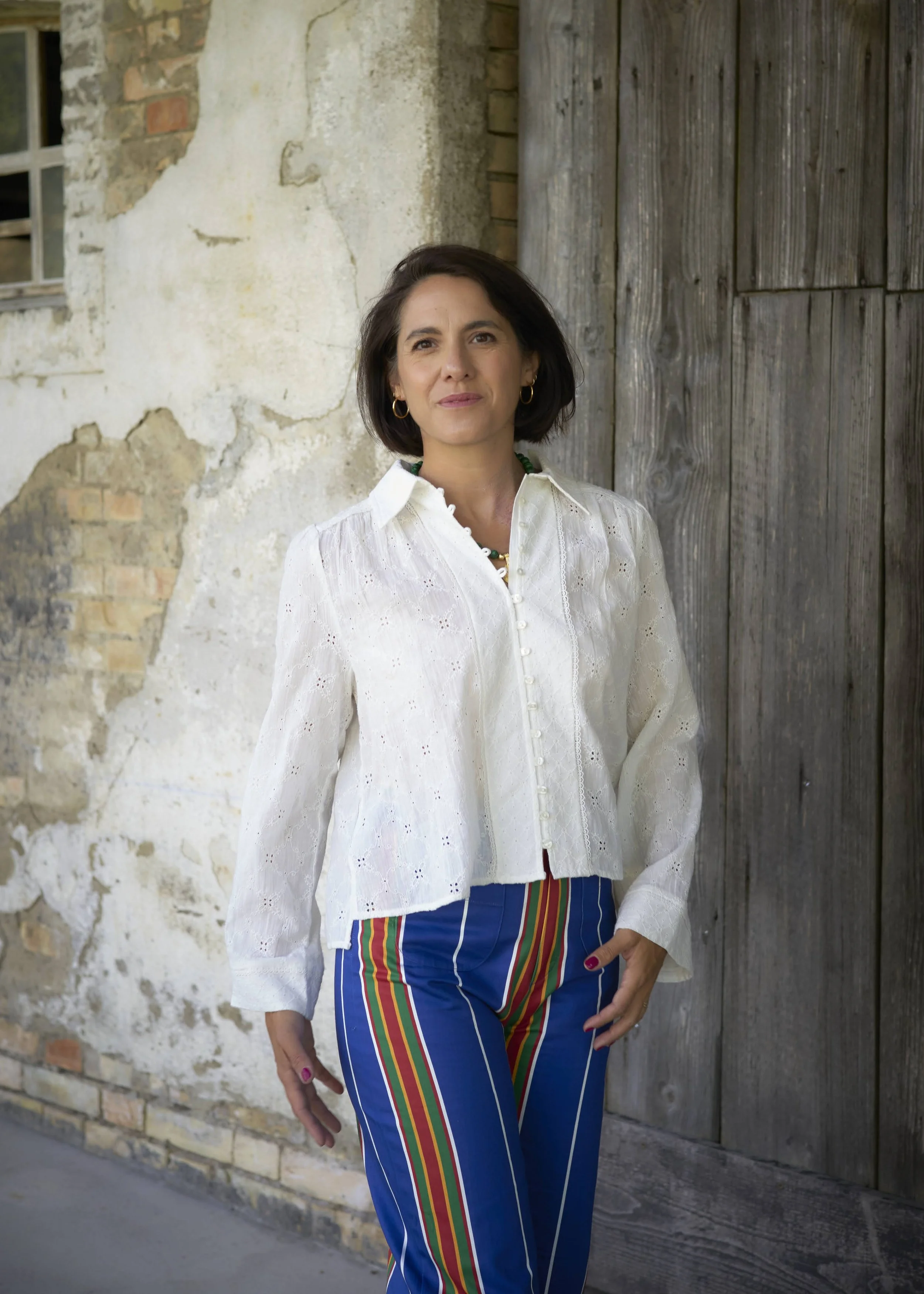 A woman with dark hair wearing a white button-up blouse with eyelet pattern and striped blue pants with rainbow stripes on the side, standing against a rustic wall with exposed bricks and weathered wooden doors.