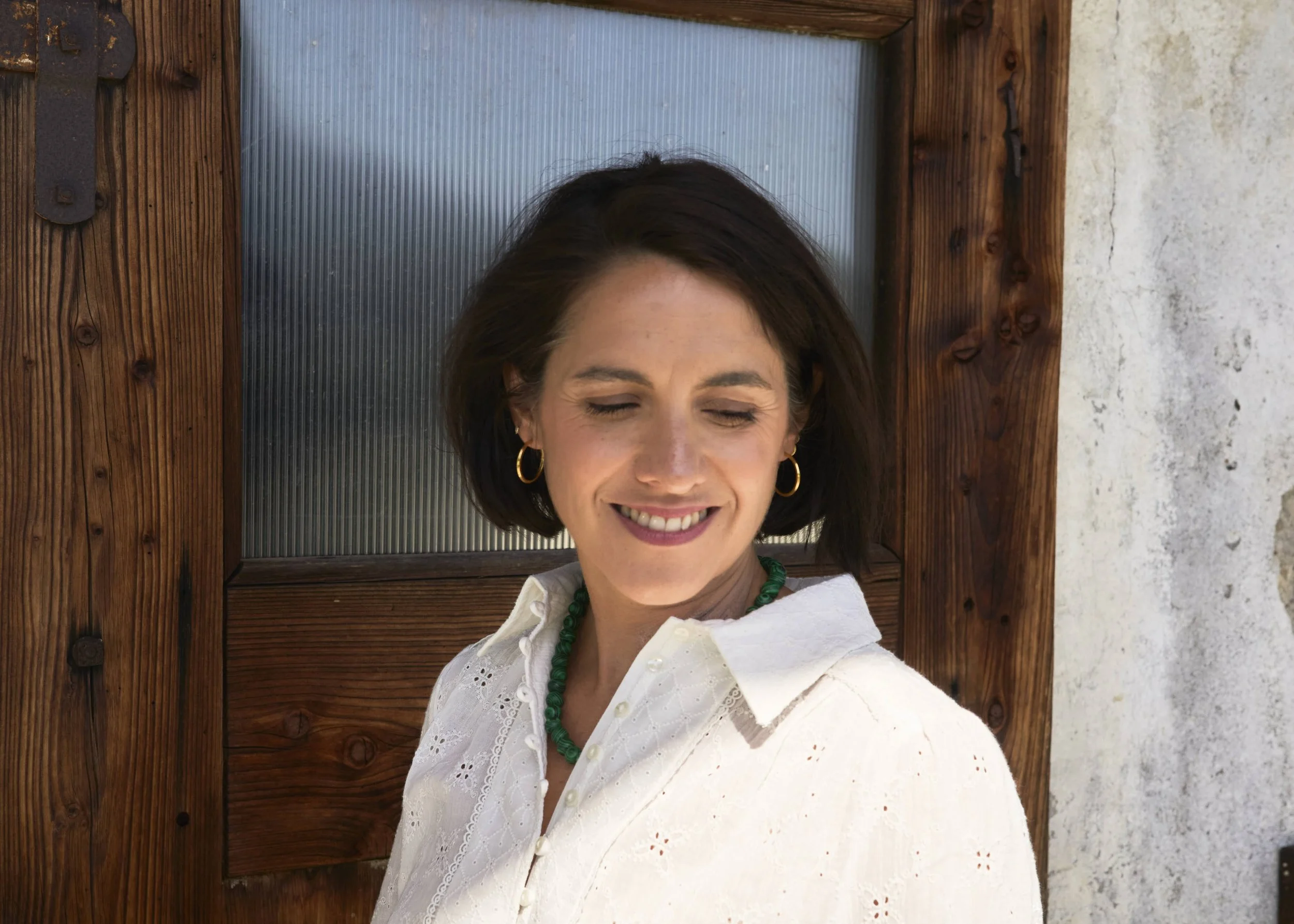 A woman with short brown hair, closed eyes, and a smile, wearing gold hoop earrings, a green beaded necklace, and a white embroidered blouse, standing in front of a wooden door with a frosted glass panel, outdoors during daytime.