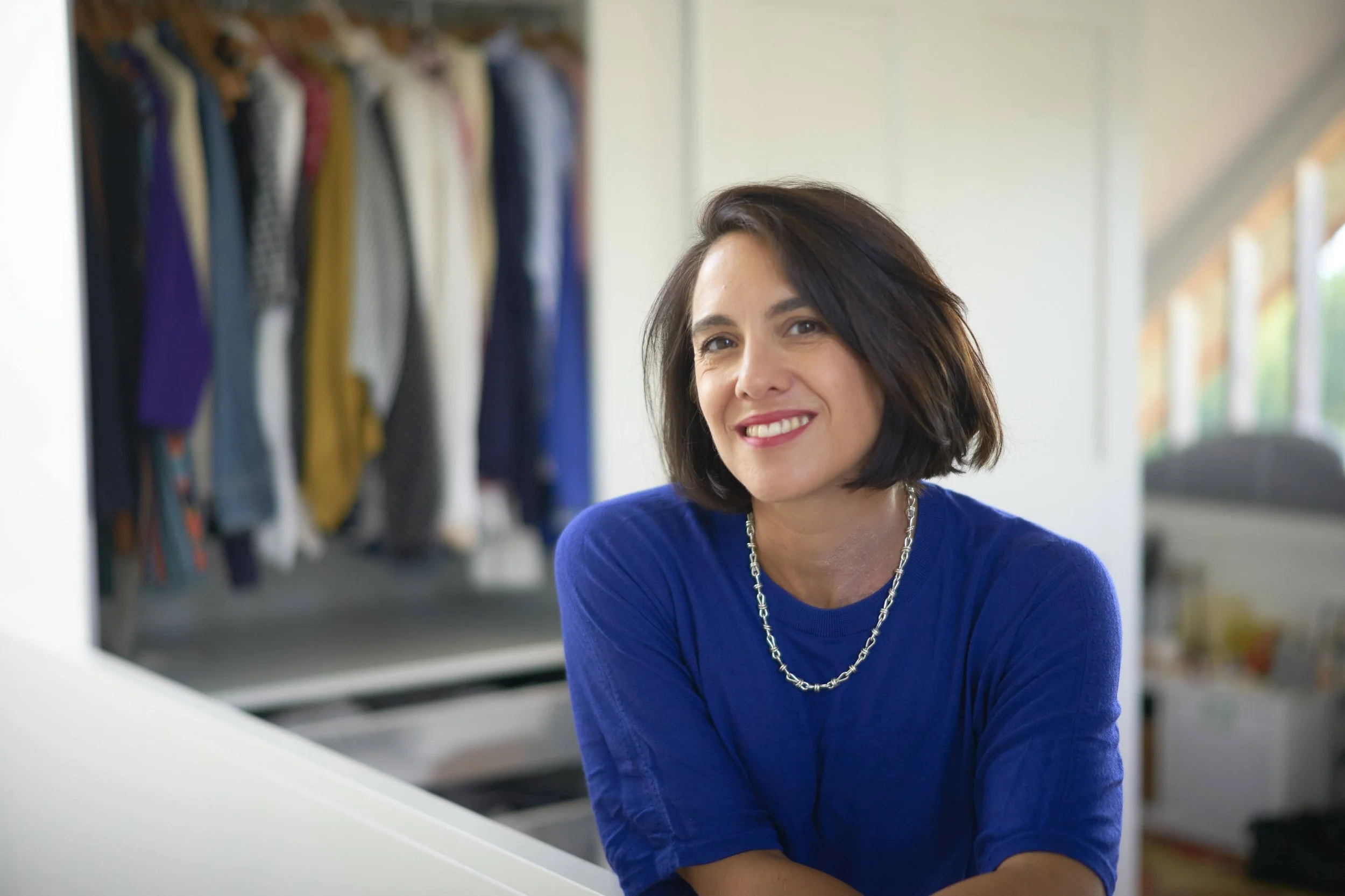 A woman with shoulder-length brown hair, wearing a blue top and a silver necklace, smiling while sitting at a piano in a room with a clothing rack in the background.