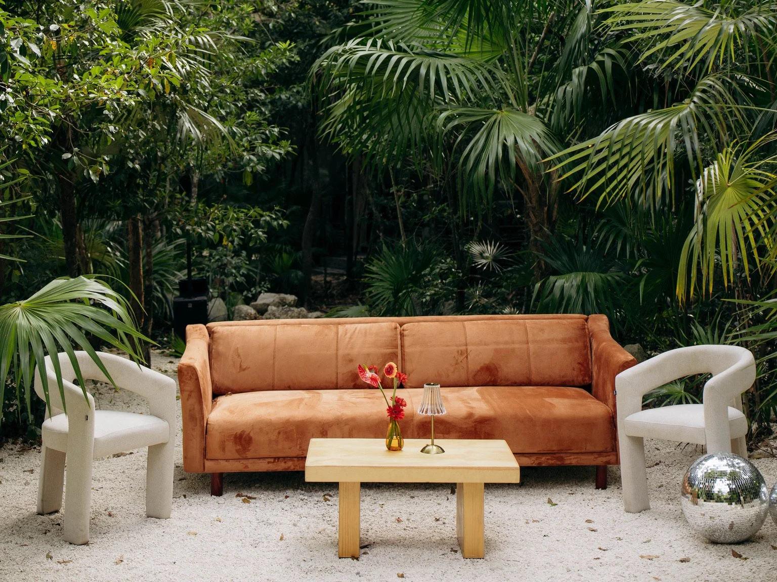 Living room set with a tan velvet sofa, white armchairs, a coffee table with a vase of red flowers, a small lamp, a mirrored ball, and a lush green jungle background.