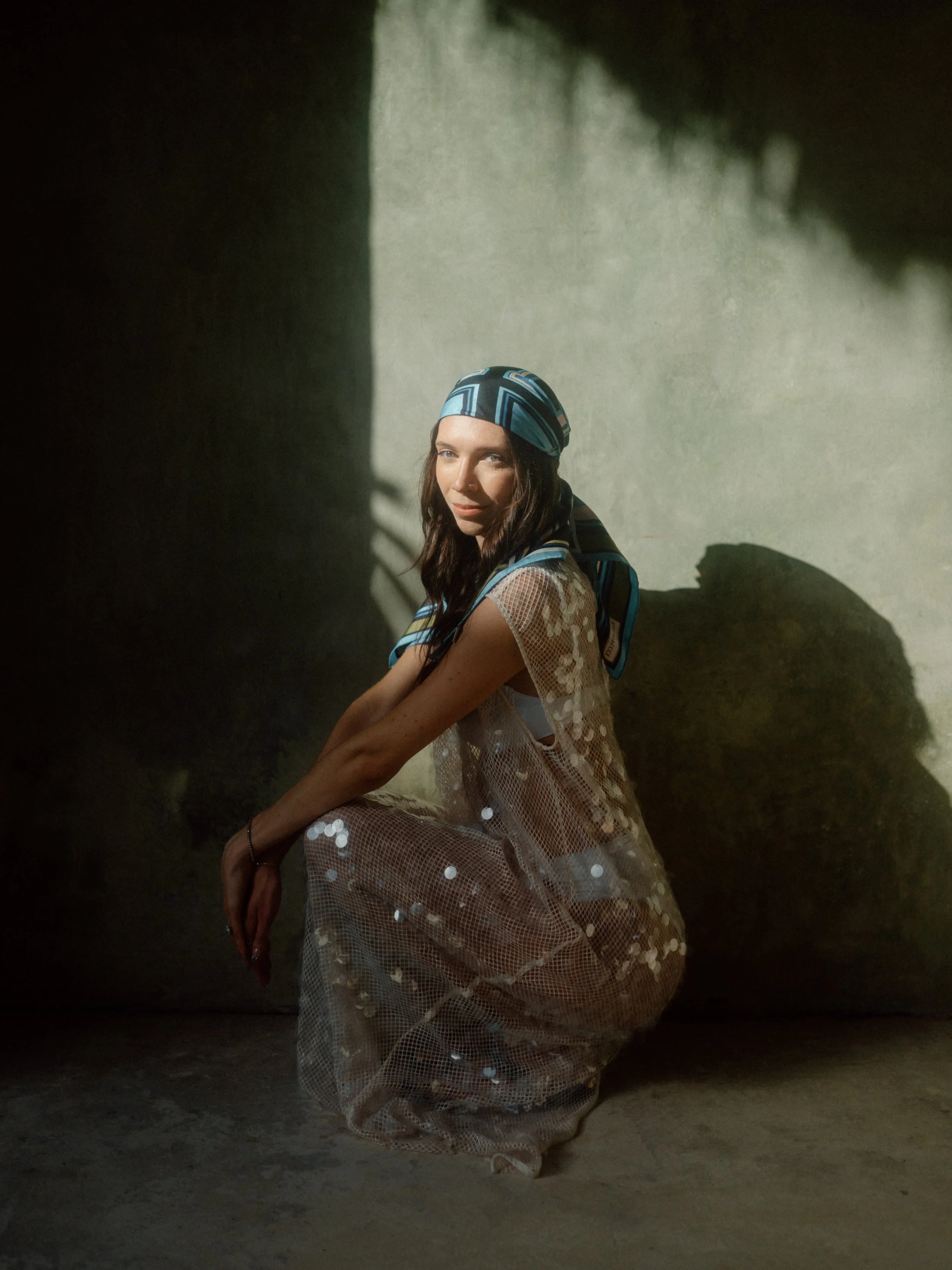 A woman with long brown hair wearing a blue patterned headscarf, a white mesh top, and a transparent dress with white polka dots, sitting on the ground with her legs crossed in a dimly lit space with a concrete wall behind her.