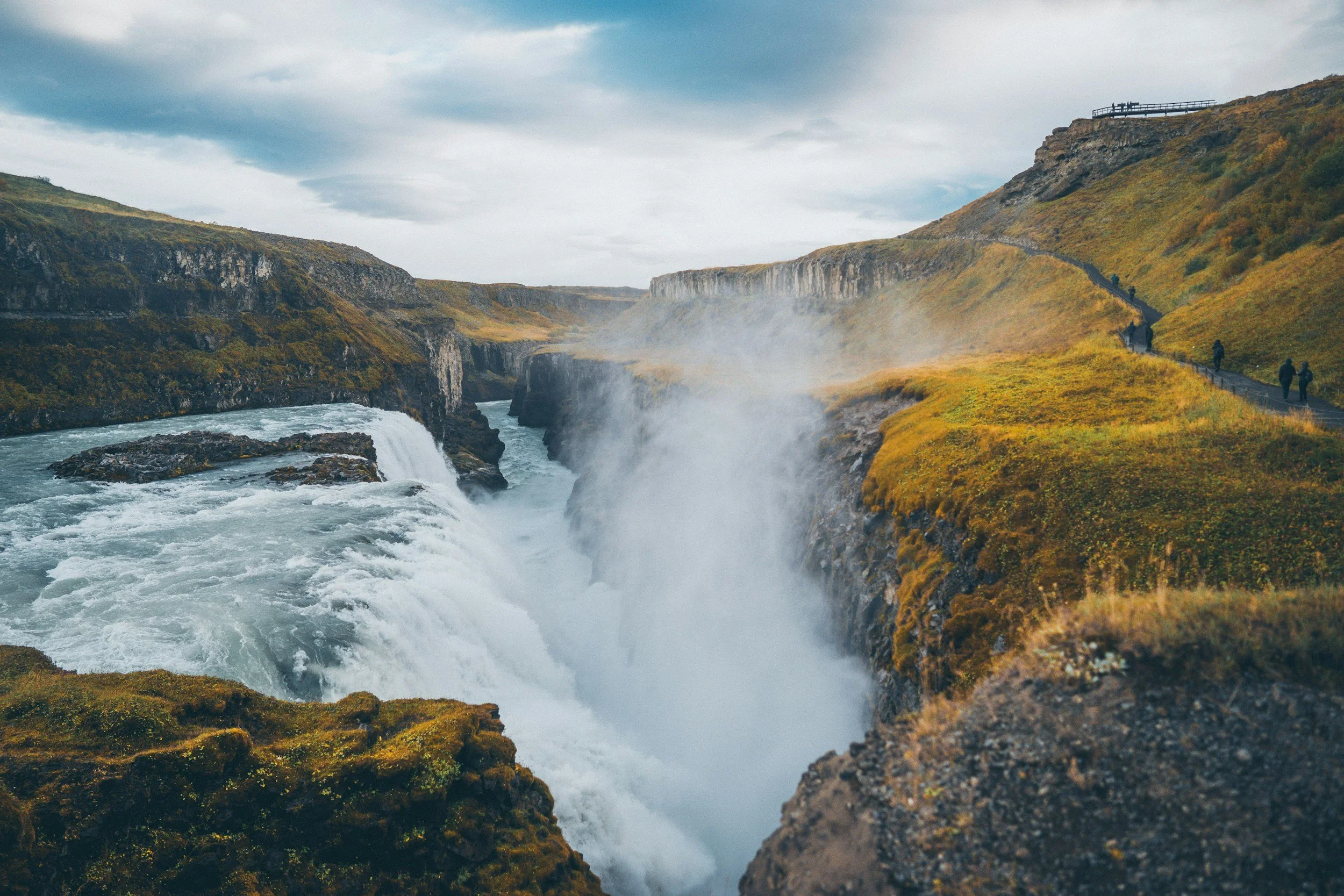 View of a deep canyon with a river flowing through the bottom, mist rising from the waterfall, and a pathway with people walking along the cliffside on the right. Overcast sky with scattered clouds.