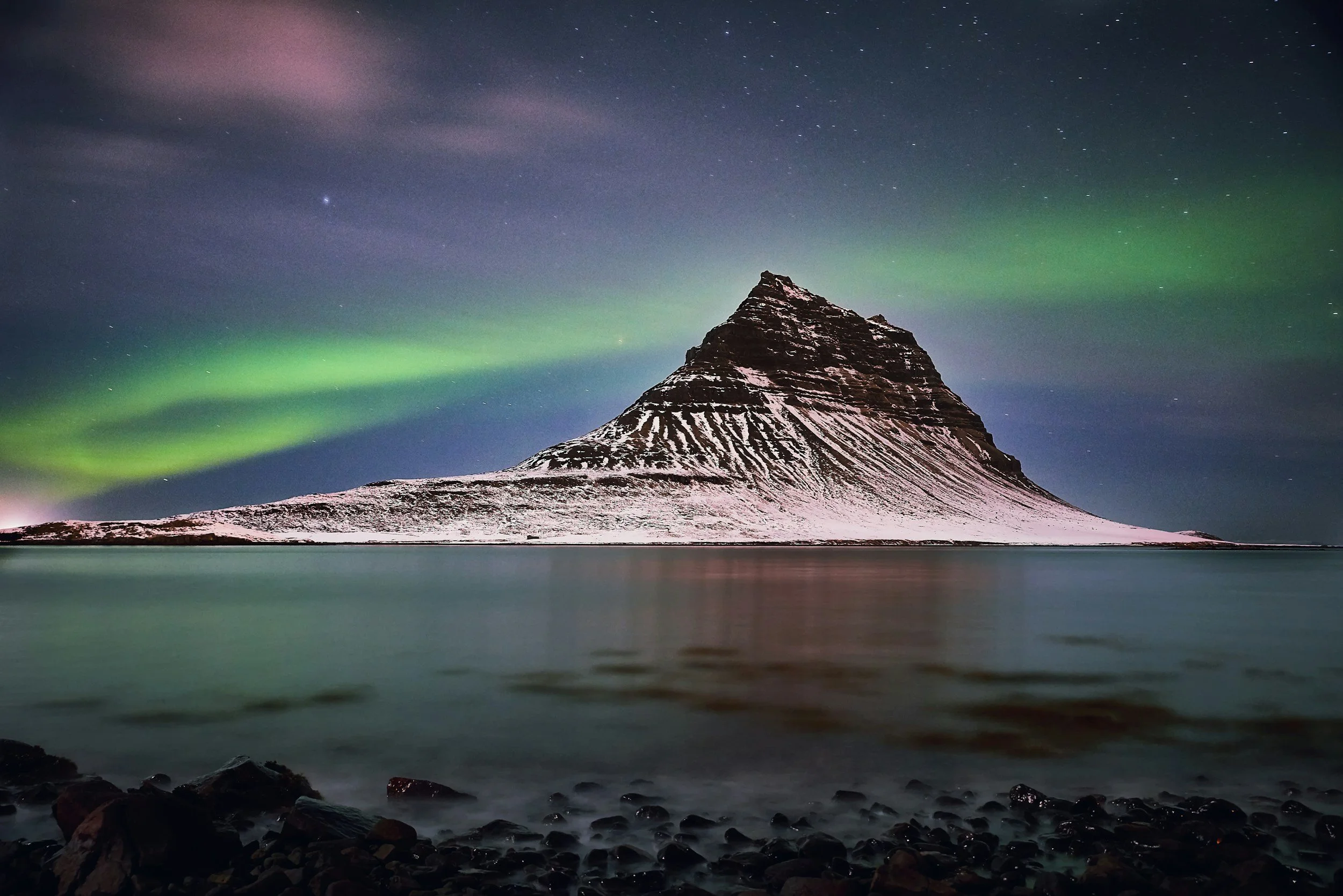 Mountain with snow and waves with rocky shoreline in front, northern lights in the night sky.
