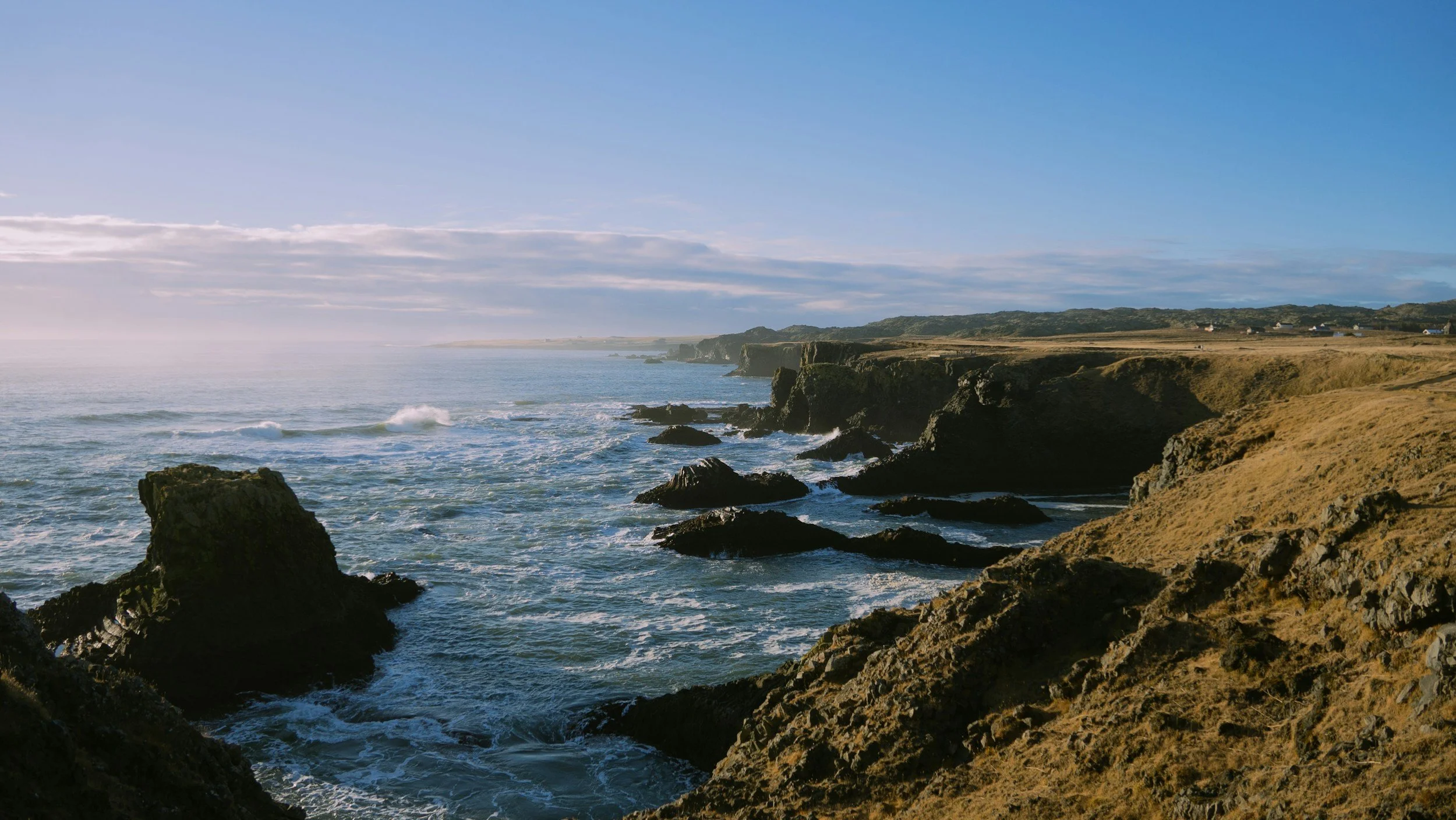 Coastal cliffs with rocks and waves crashing in the ocean, under a partly cloudy sky, with some houses on the distant land.