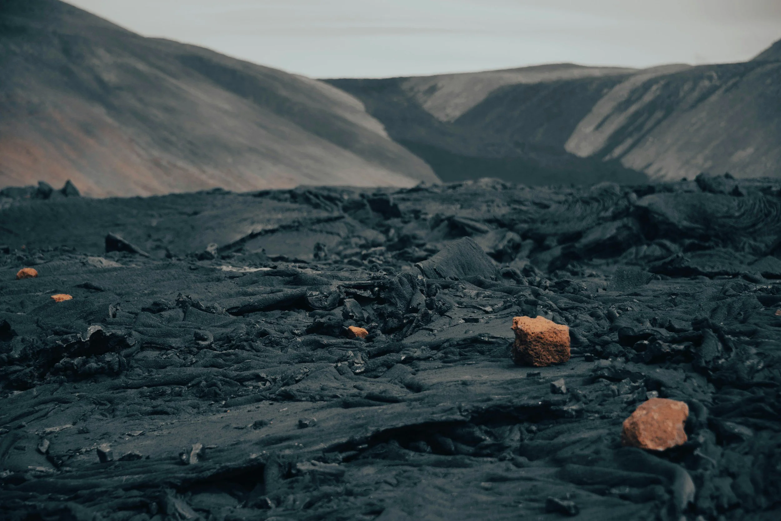 A dark, rocky landscape with volcanic rocks in the foreground and rolling hills or mountains in the background, under a cloudy sky. Volcanic rocks and stagnant lava near Grindavik in Iceland.