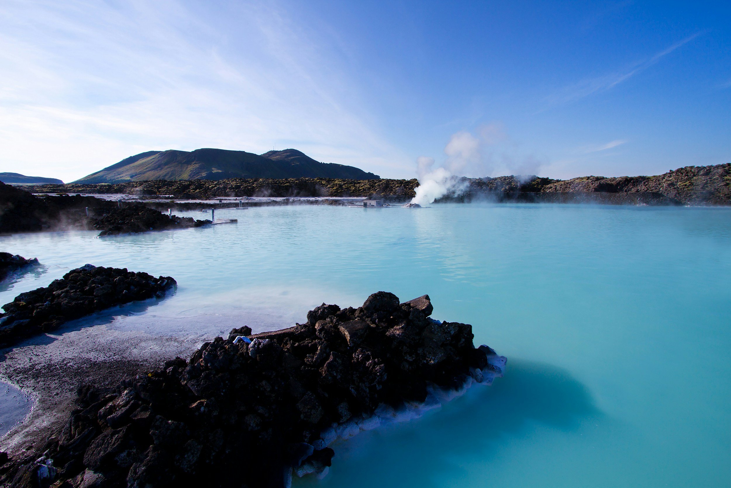 A geothermal hot spring with steam rising from the waters of Blue Lagoon, surrounded by volcanic rocks with a mountain in the background under a blue sky.