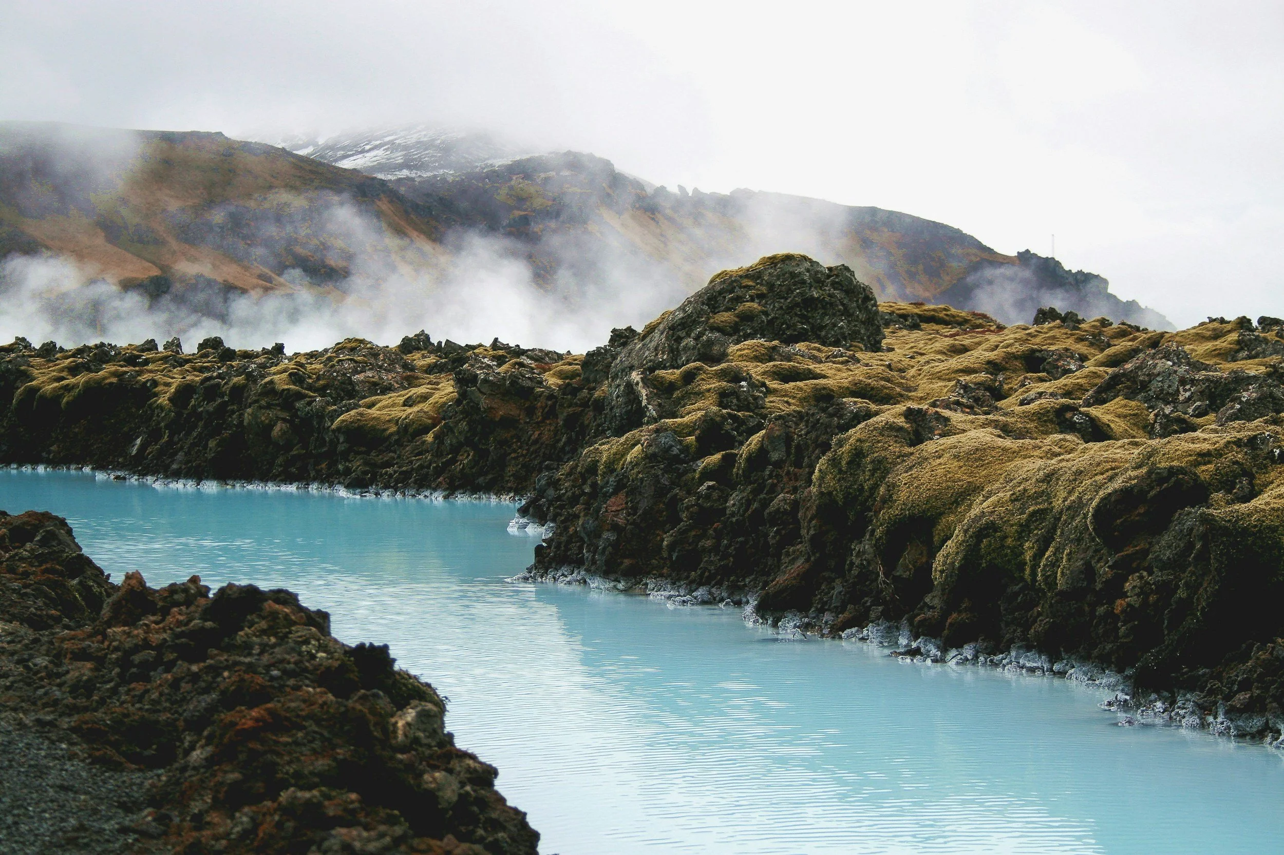Volcanic landscape with moss-covered rocks at Blue Lagoon, steaming vents, and a geothermal hot spring or pool beneath cloudy, foggy sky.