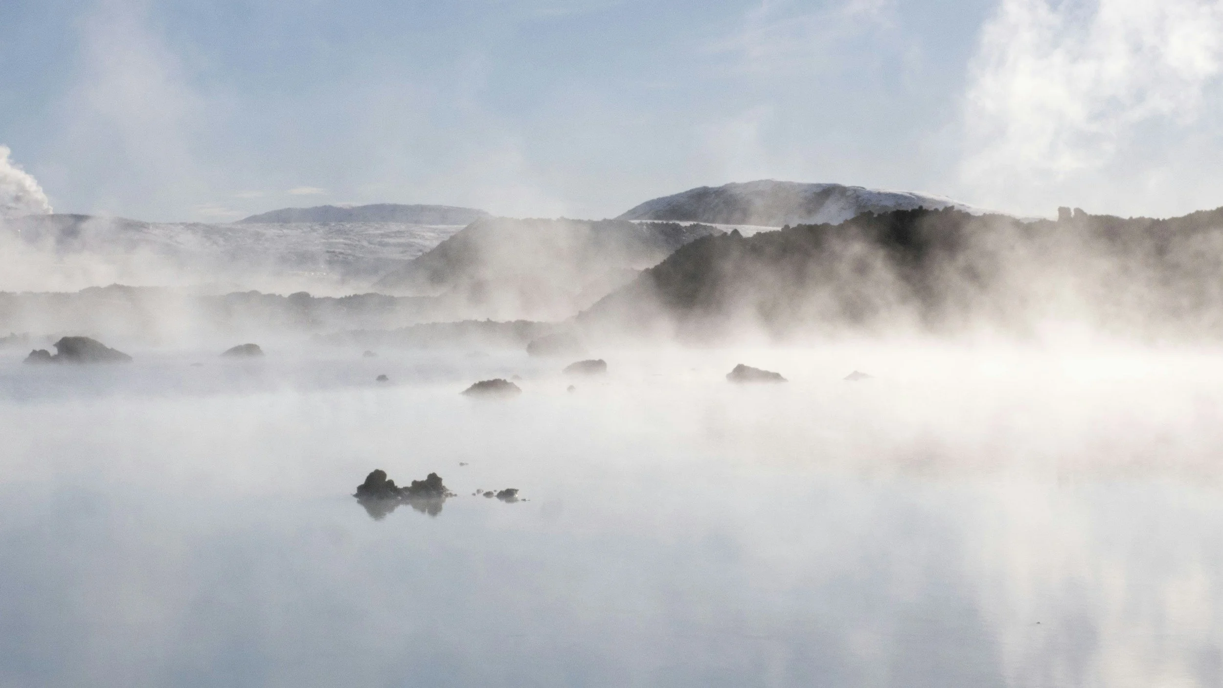 A landscape featuring snow-capped mountains near Hvammsvik with a cloudy sky, misty fog over a calm body of water with rocks protruding.