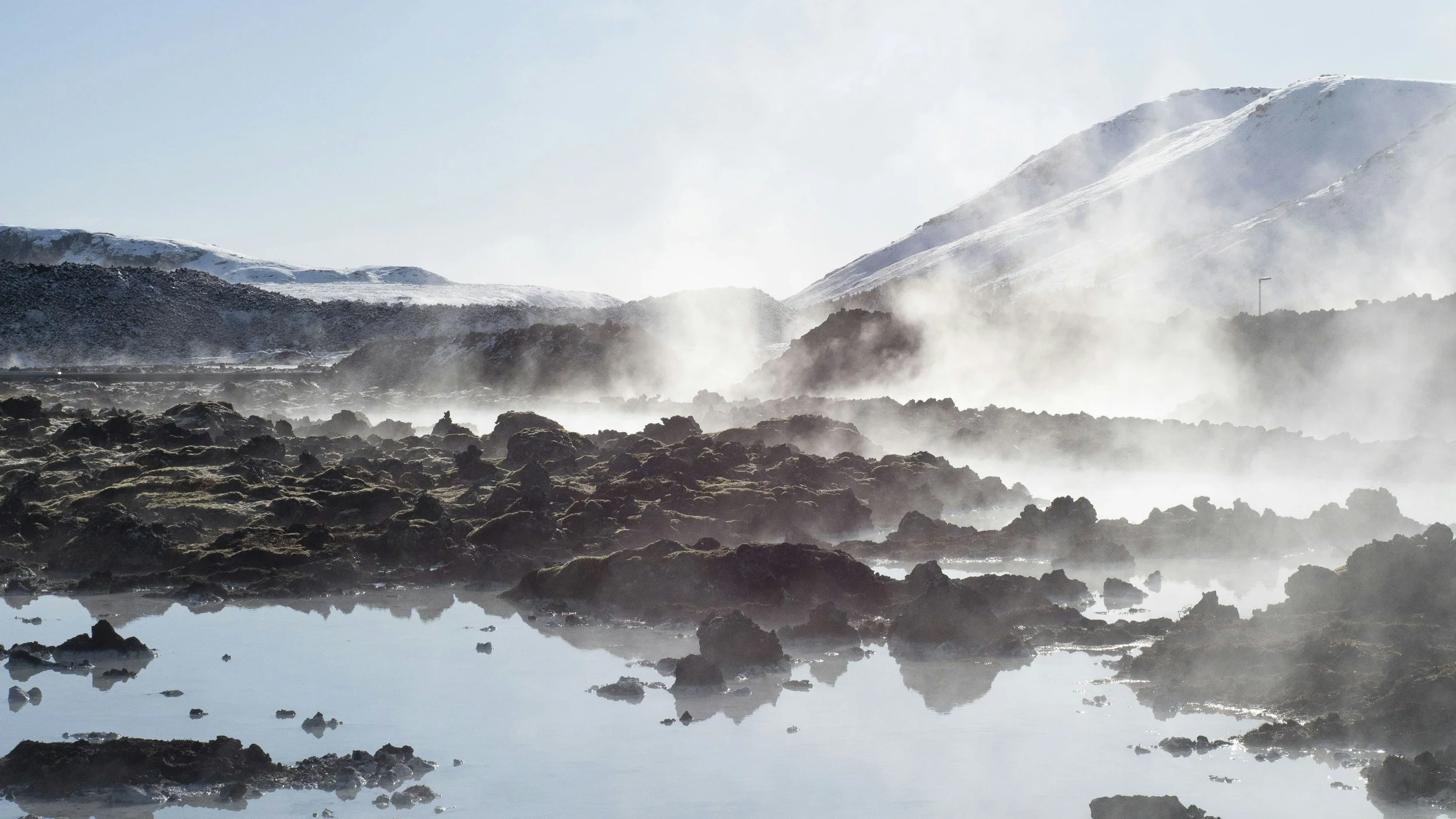 Steam rising from a rocky hot spring Hvammsvik with snow-covered mountains in the background and a partly cloudy sky.